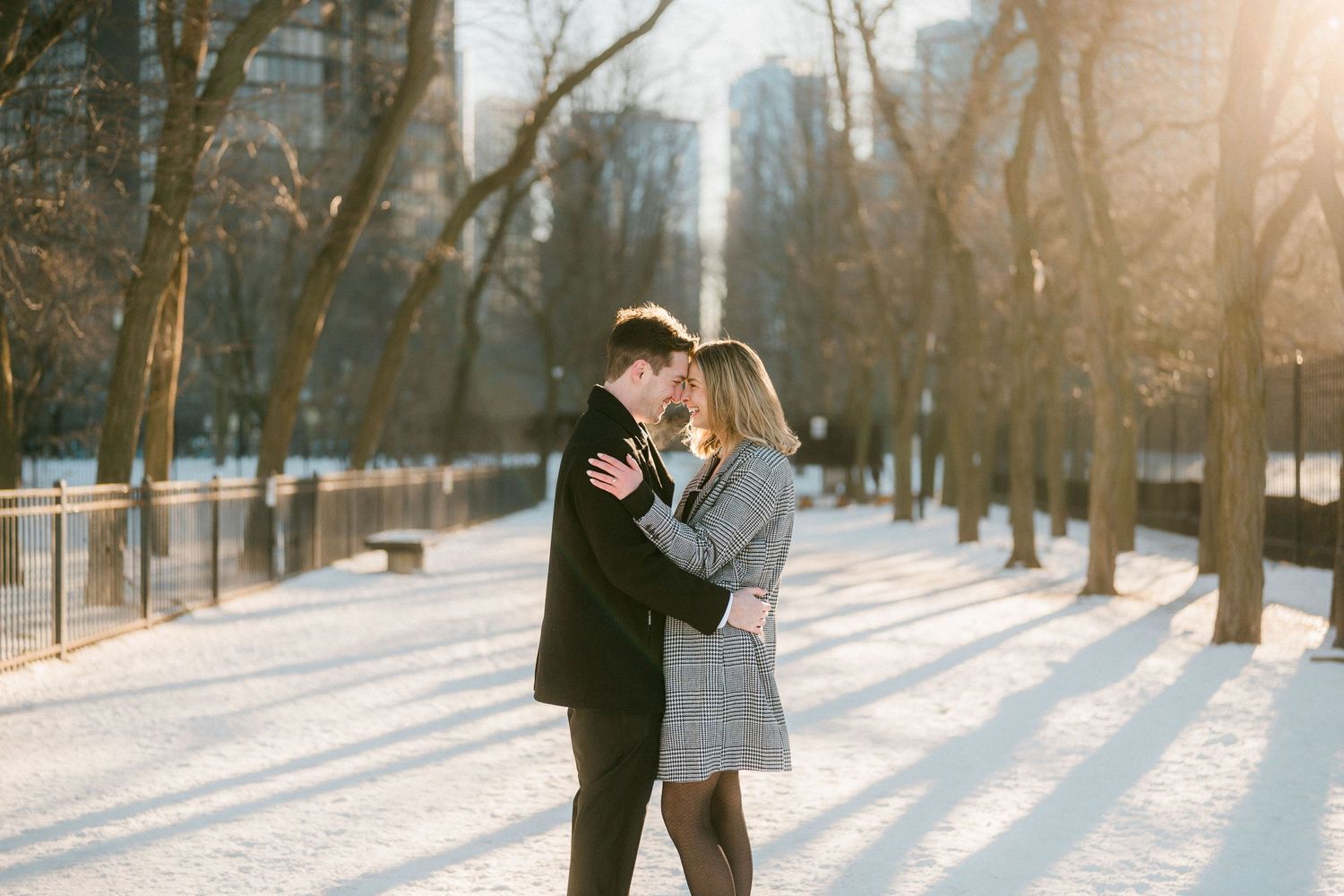 Couple embracing in snowy park with bare trees and golden sunlight in winter.