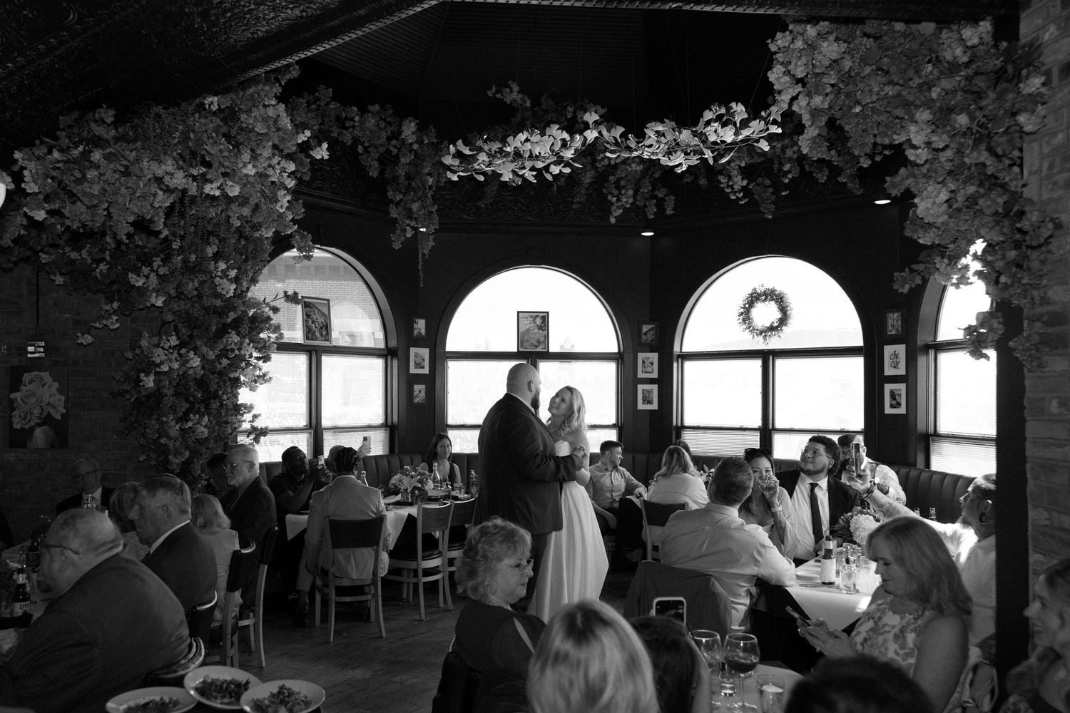 Black and white wedding reception photo with couple standing at head of room while guests dine at decorated tables.