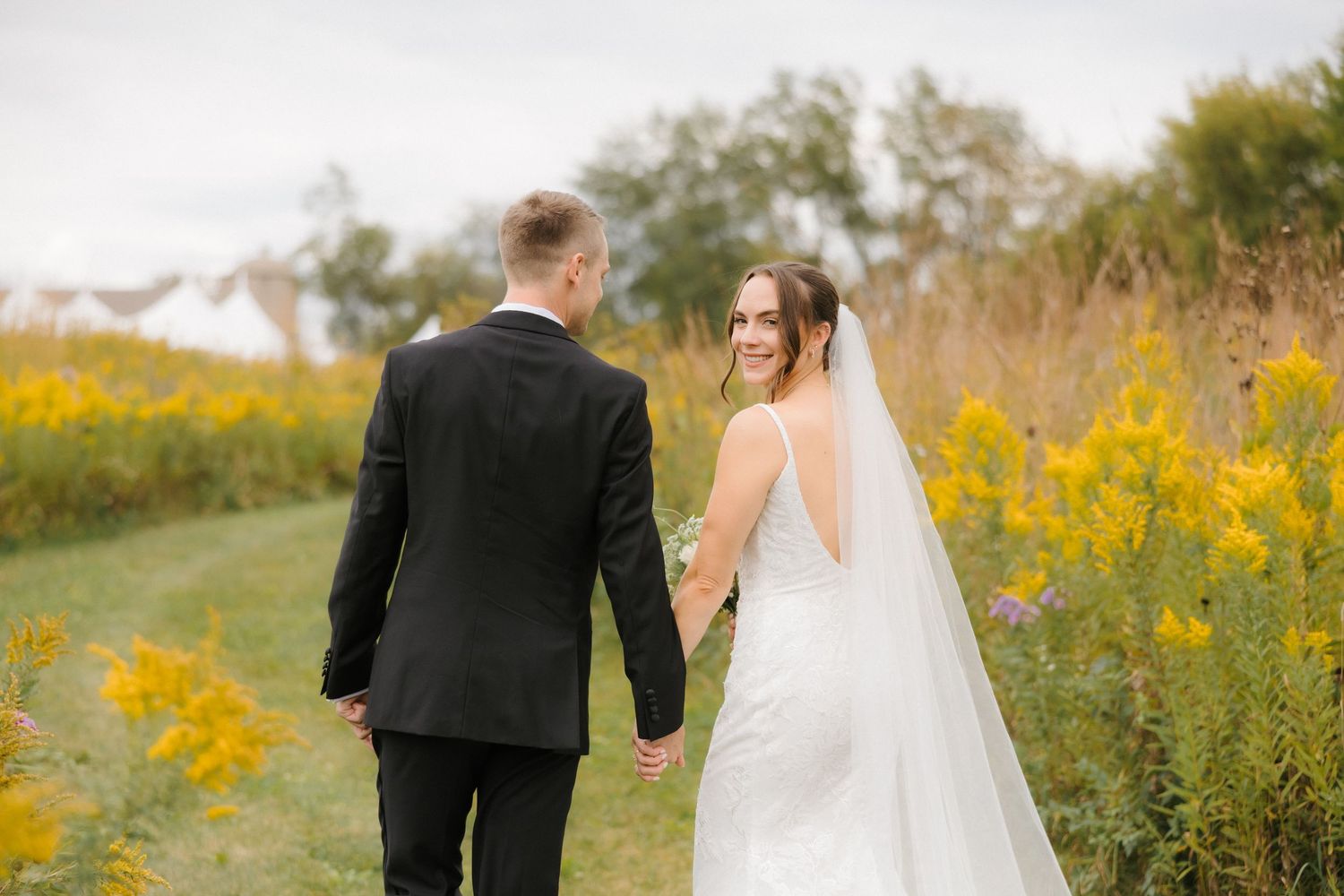 Bride in white gown and groom in tuxedo walking hand in hand through yellow wildflower field.