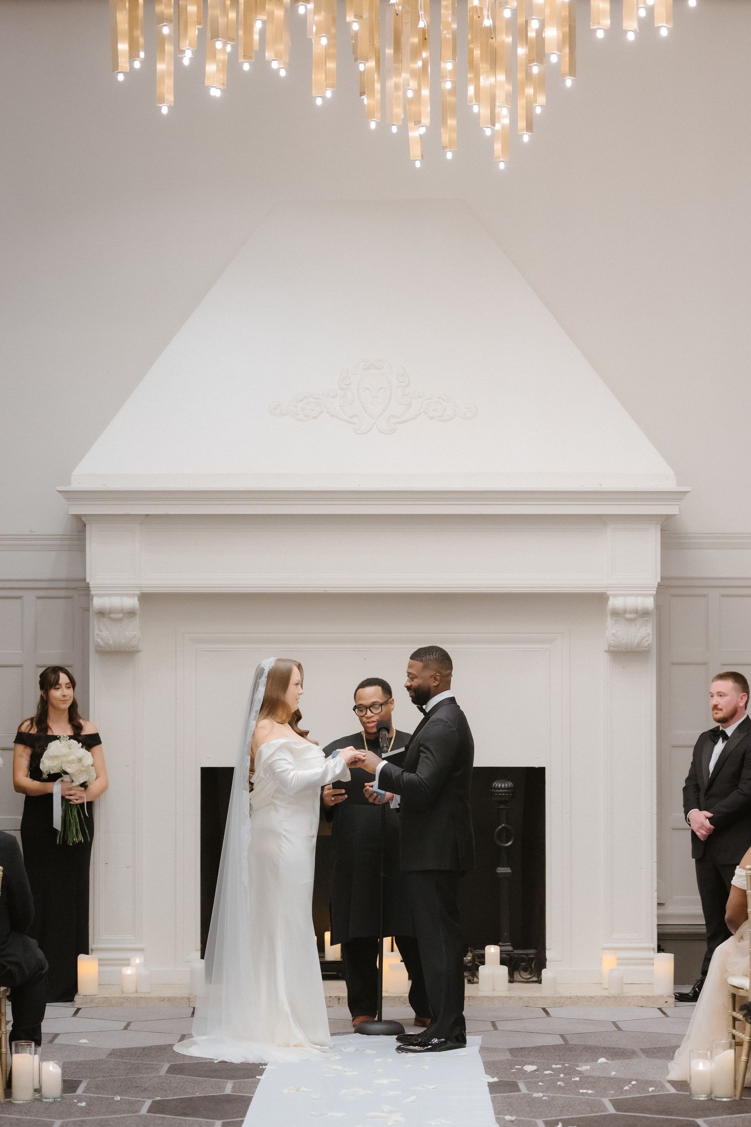 Bride and groom exchange vows before an officiant at an elegant indoor wedding ceremony with a white fireplace backdrop.