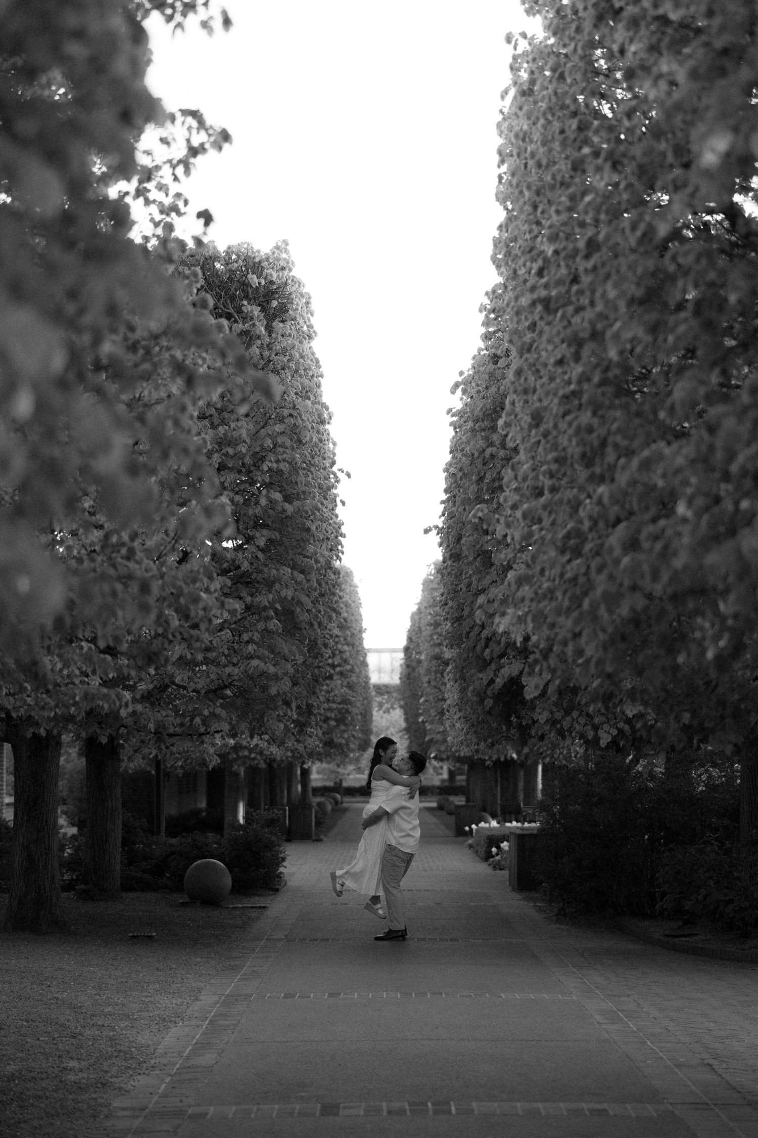 Black and white photo of a woman twirling in a tree-lined pathway, wearing a flowing dress.