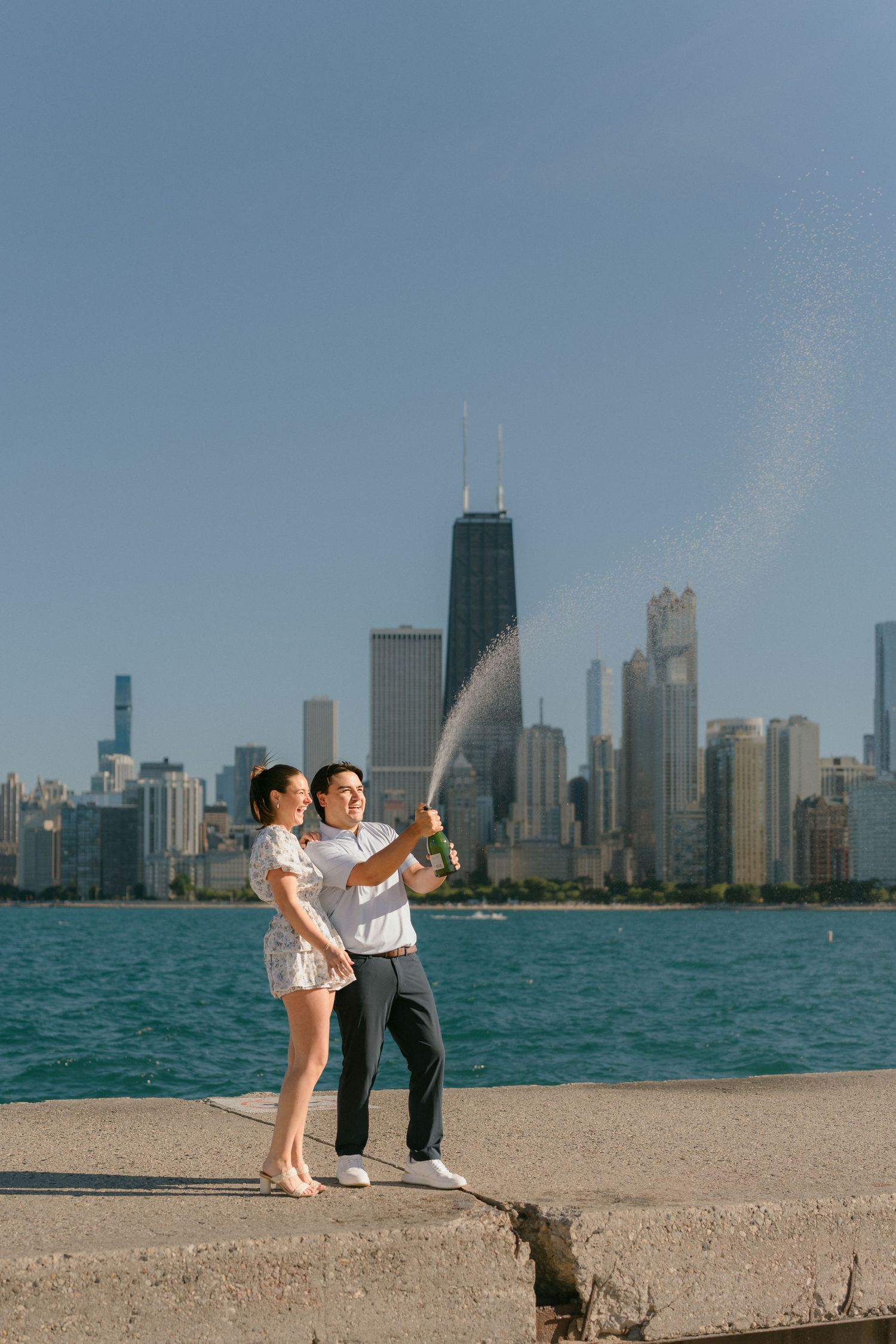 Couple popping champagne bottle on lakefront with stunning Chicago city skyline behind them.
