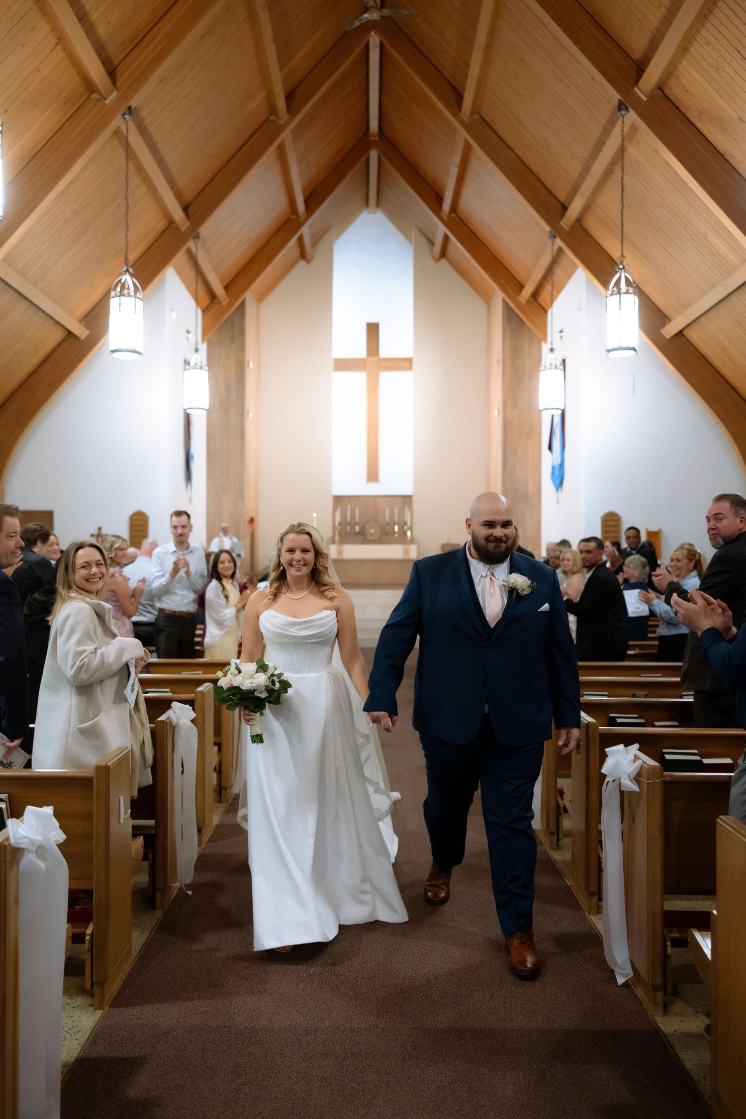 Bride and groom walk down church aisle after wedding ceremony, guests applauding, wooden chapel with cross backdrop.