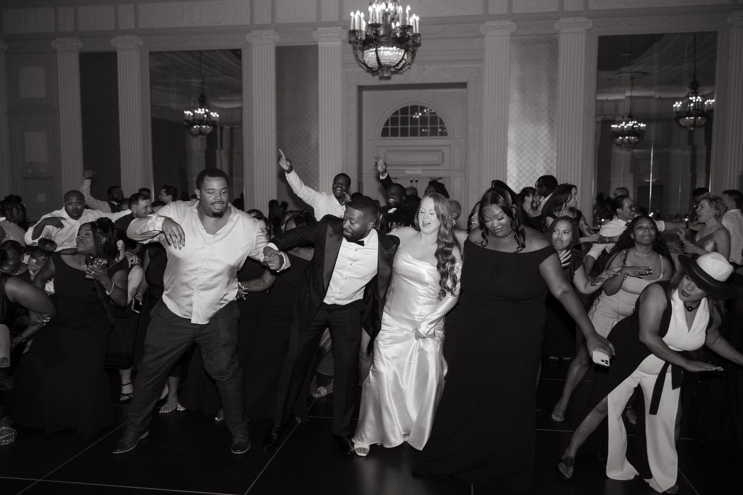 Black and white photo of wedding guests dancing joyfully at a elegant ballroom reception.