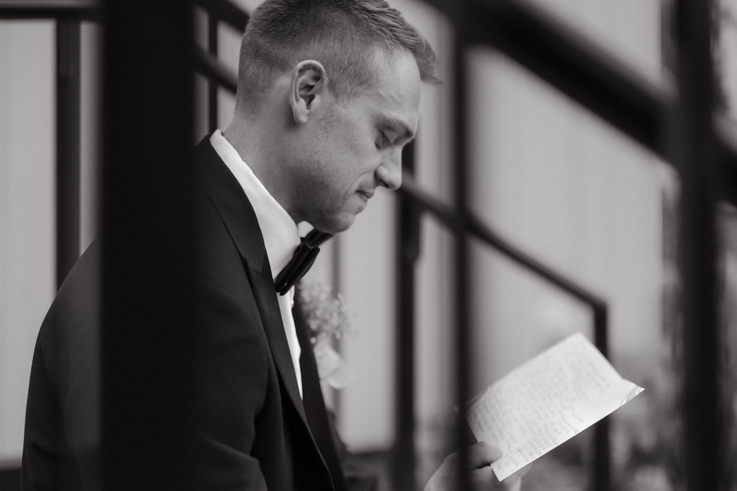 Black and white photo of groom in tuxedo reading letter before wedding ceremony.