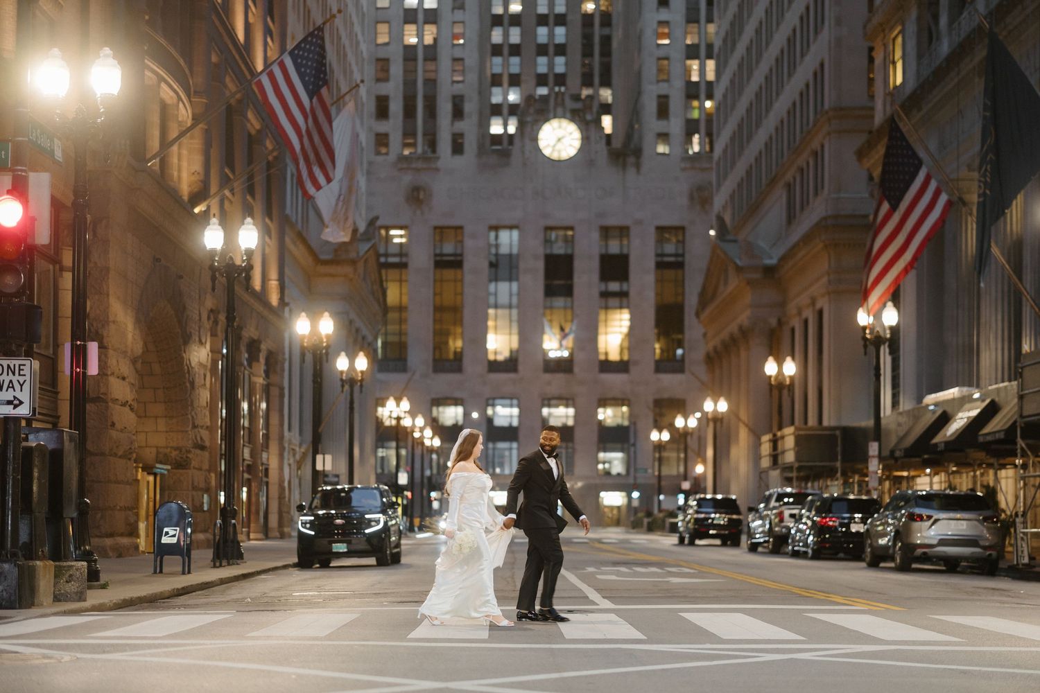 Bride and groom hold hands crossing a Chicago street at night with illuminated buildings and American flags.
