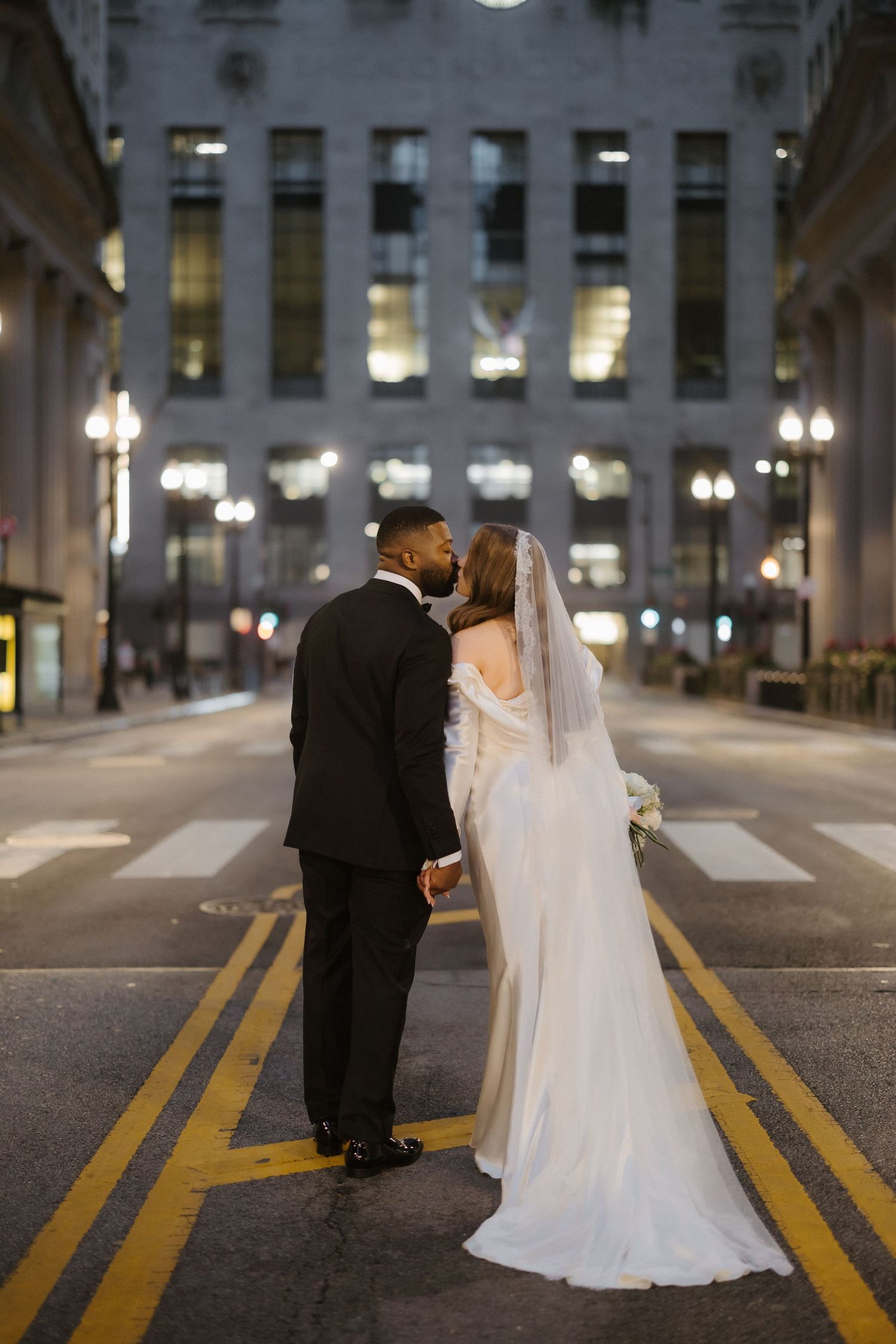 Bride and groom share a kiss on a city street at night, with a grand illuminated building backdrop.