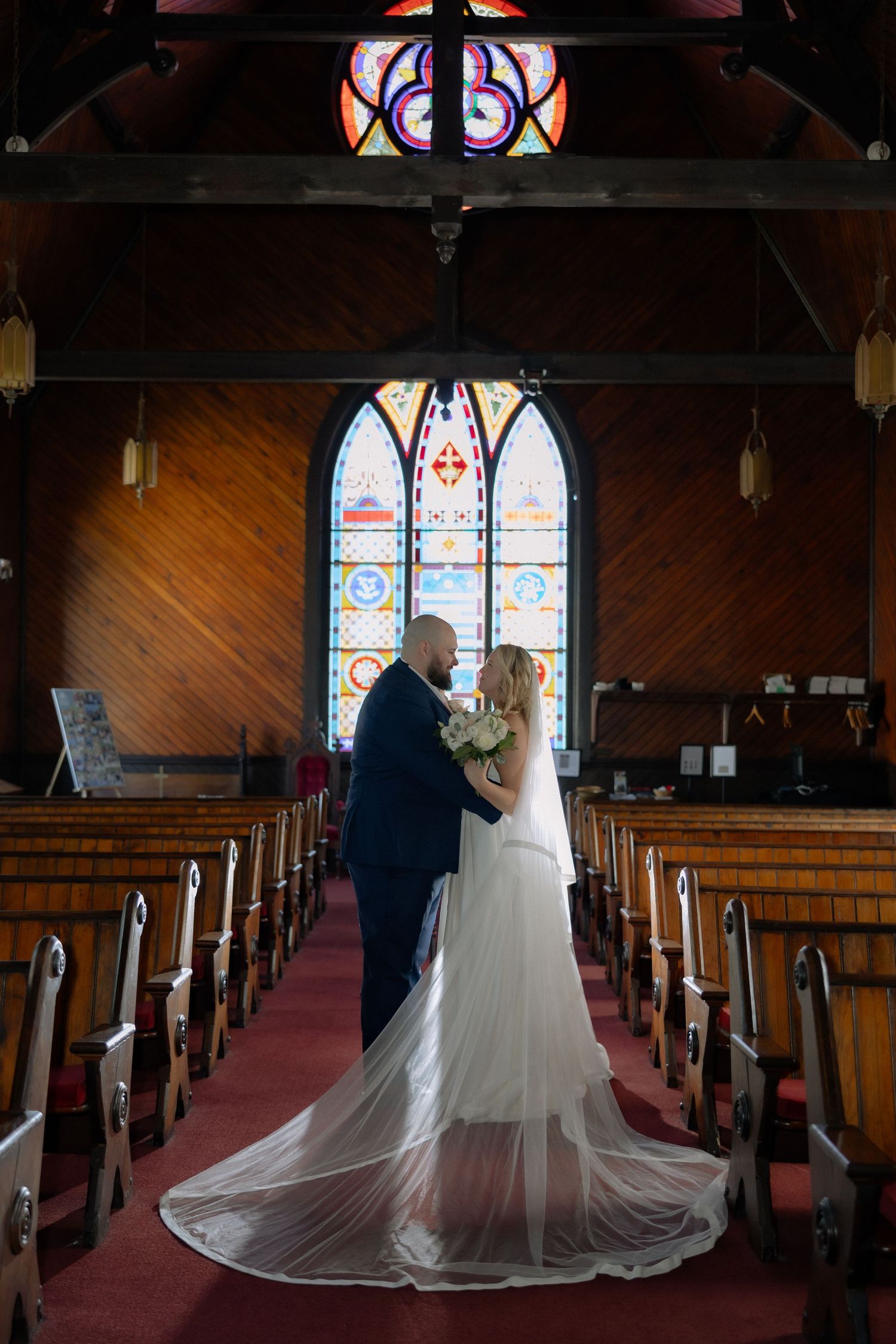 Bride and groom share a romantic moment at the altar of a wooden chapel with stunning stained glass windows.