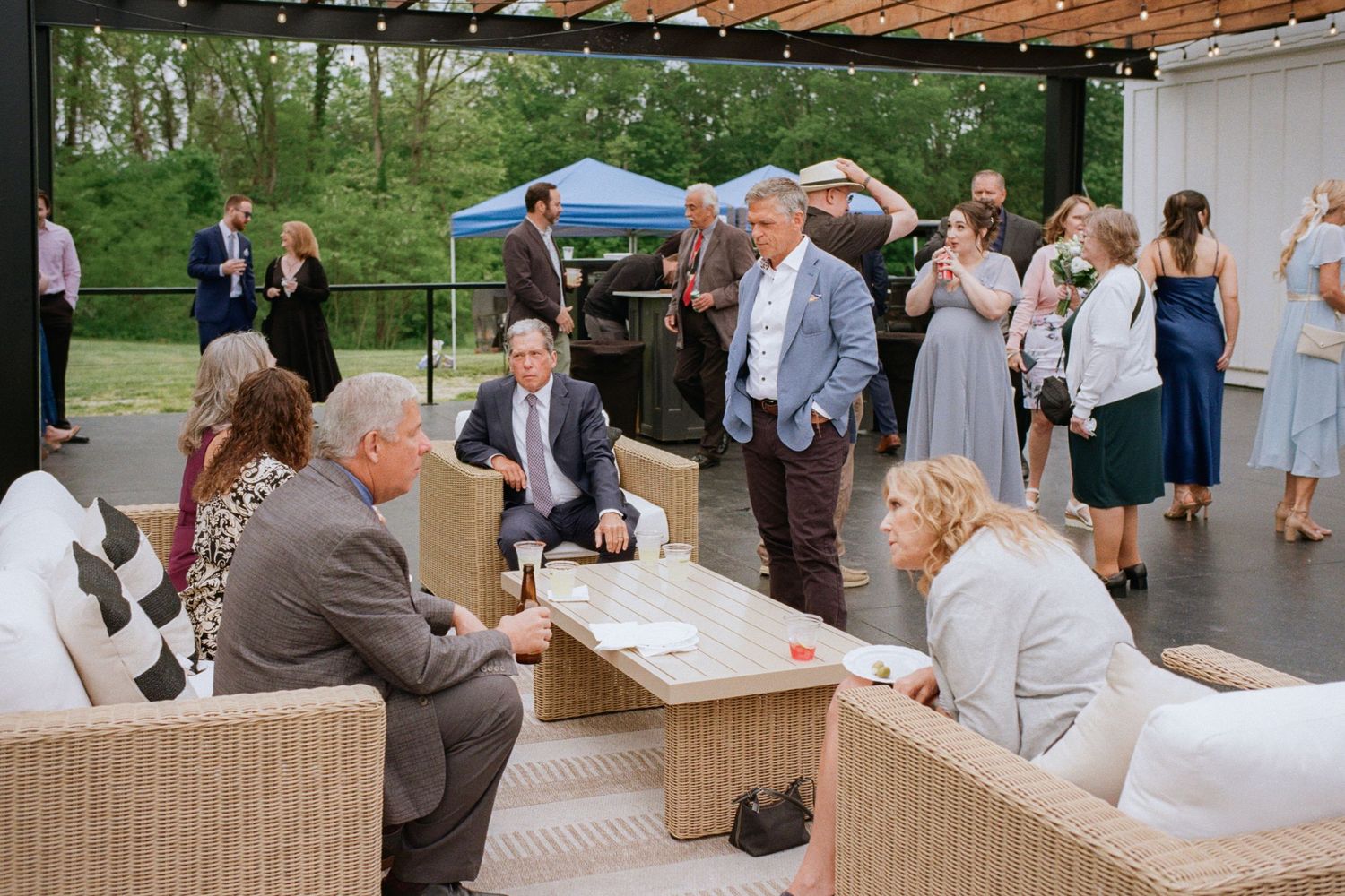 Guests mingle and relax on wicker furniture at an outdoor wedding cocktail hour on a covered patio.