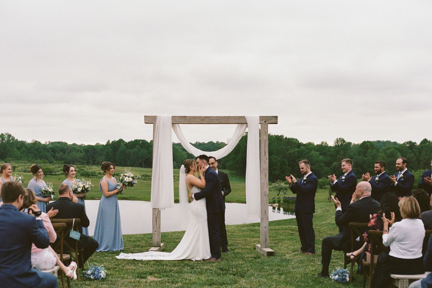Outdoor wedding ceremony with couple kissing under rustic wooden arch draped in white fabric, surrounded by guests on a green farm.