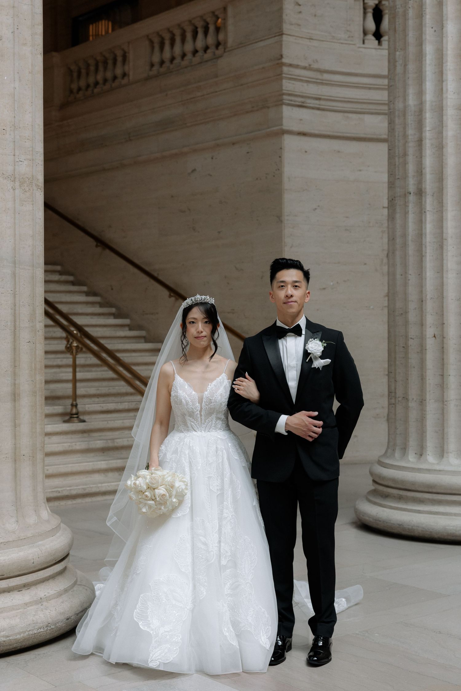 Elegant bride and groom pose between classical columns at grand architectural wedding venue.