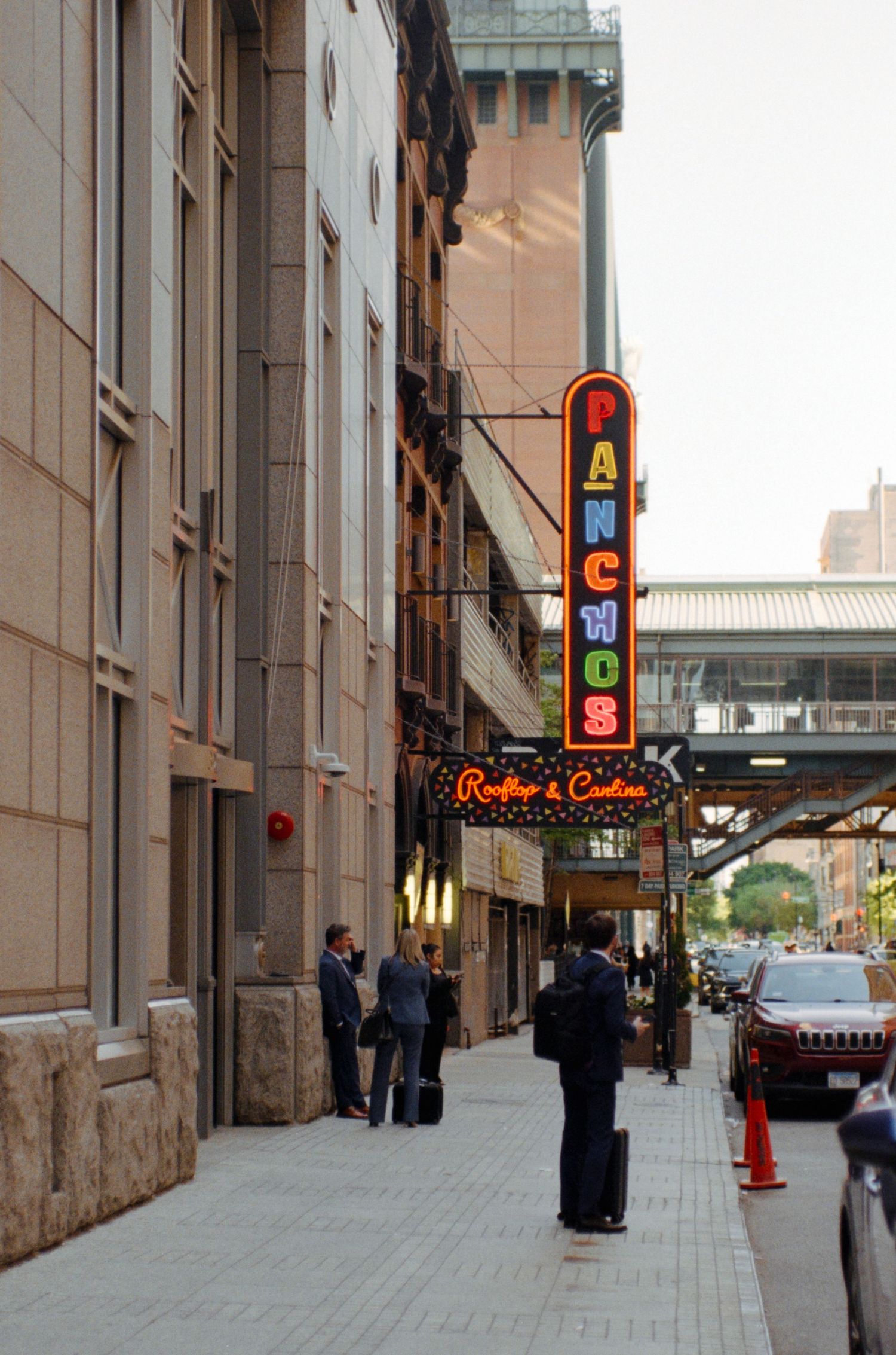 People walking past Panchos Rooftop and Cantina neon sign on a city sidewalk with tall buildings.