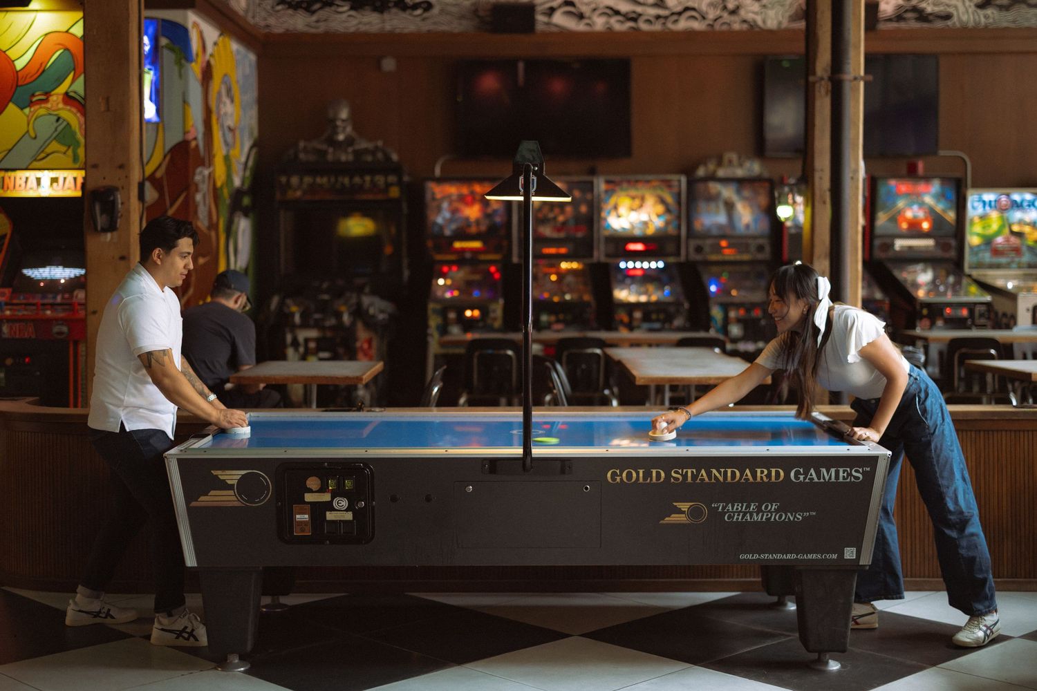 Two people playing air hockey on a Gold Standard Games table in a retro arcade bar with pinball machines in background.