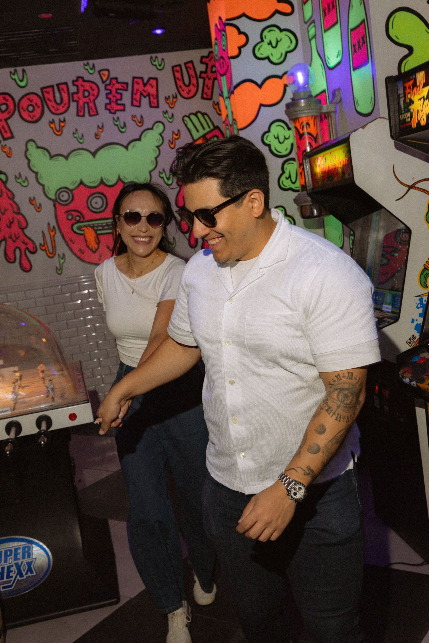 Couple in matching white shirts and sunglasses laughing at colorful arcade game venue.