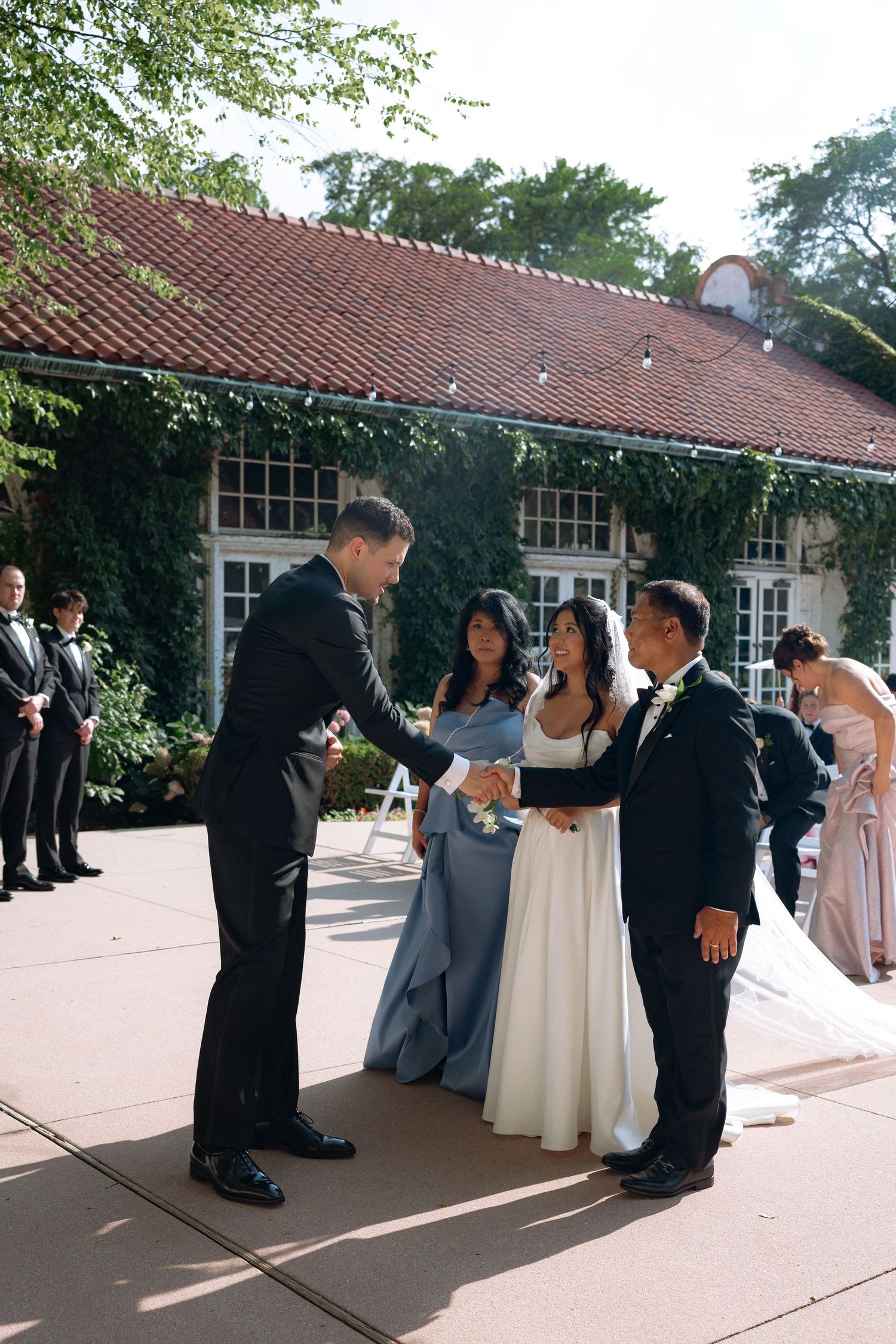 Groom shaking hands with father of the bride at outdoor wedding ceremony, with bridesmaids in blue dresses nearby.