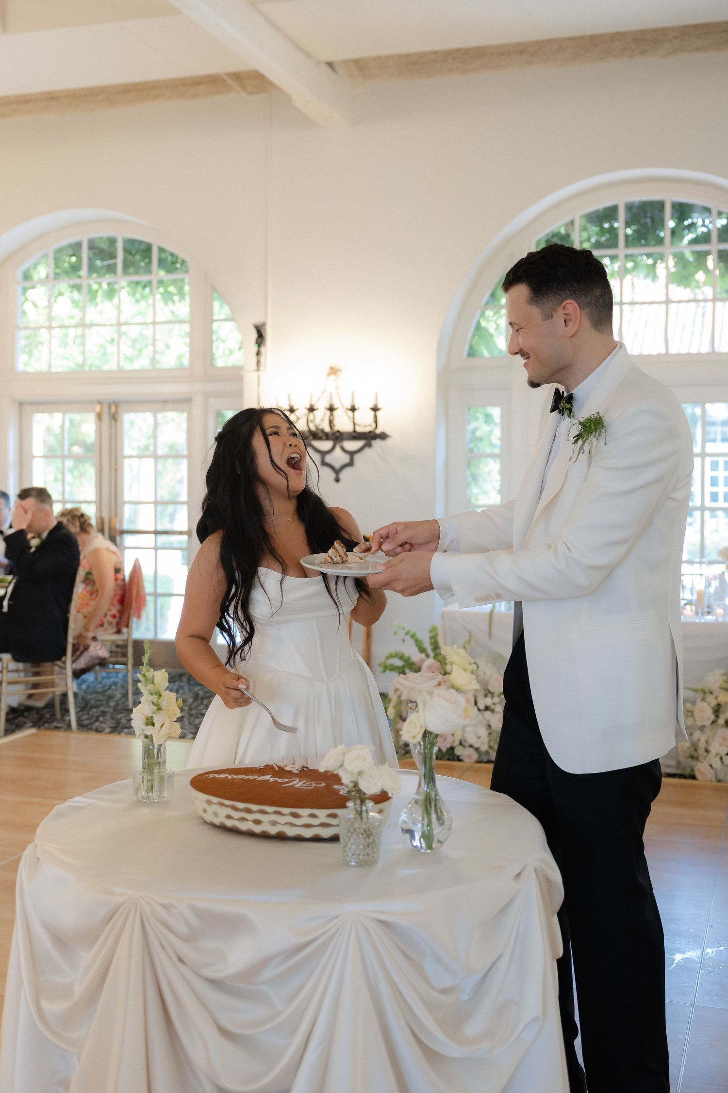 Laughing bride and groom cut their wedding pie at a decorated table in a bright reception hall.