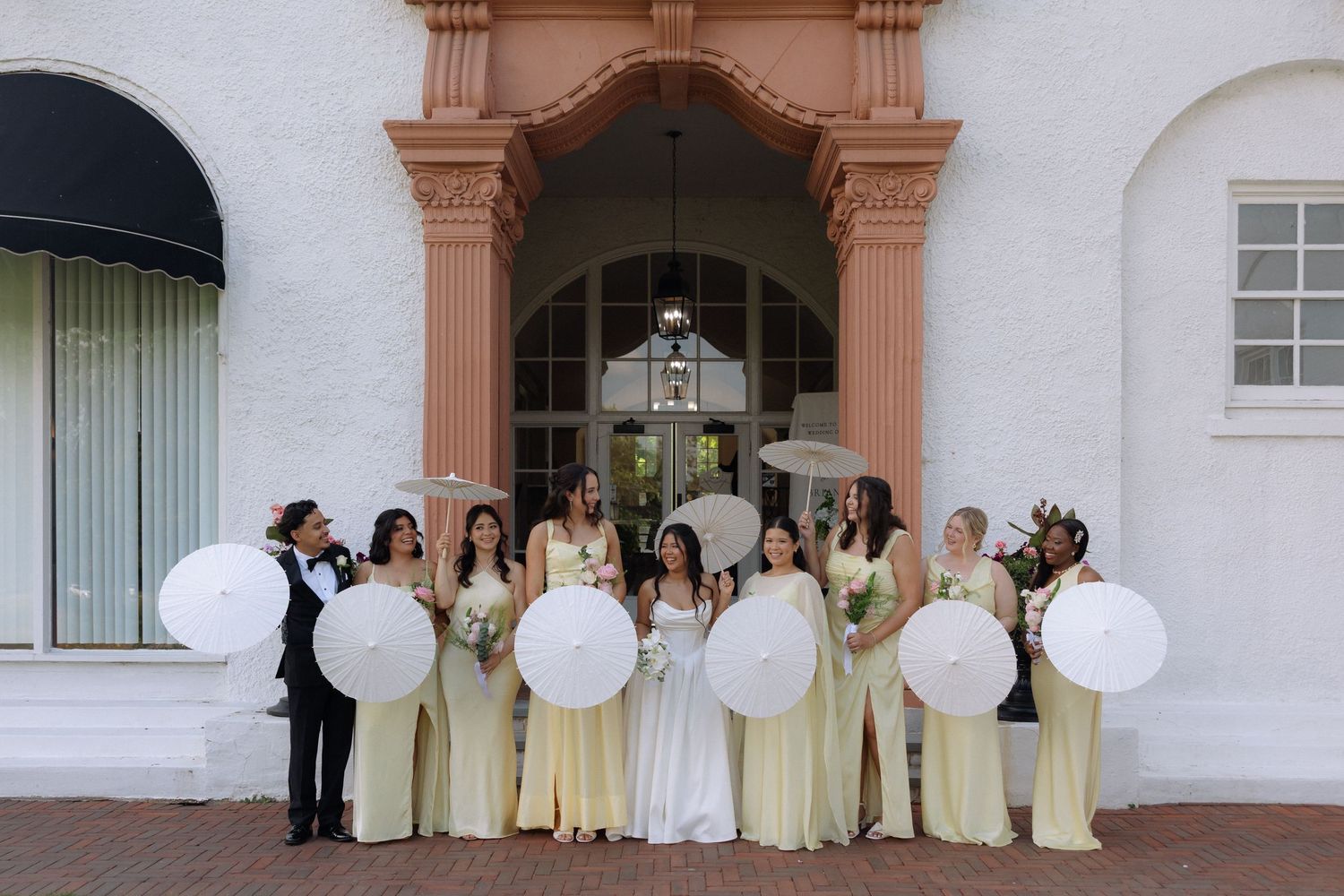 Wedding party with bridesmaids in yellow dresses holding white parasols outside elegant venue entrance.