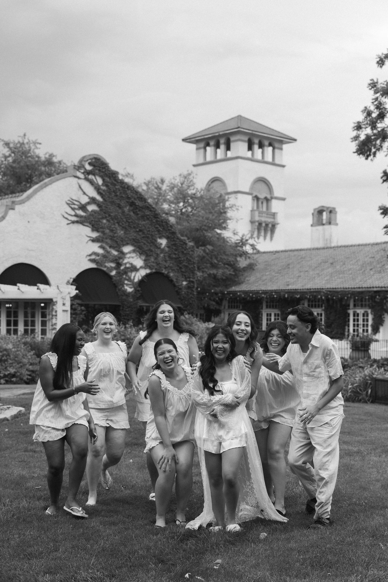 Black and white photo of a joyful group of women in white outfits laughing and running on a lawn outside an elegant venue.