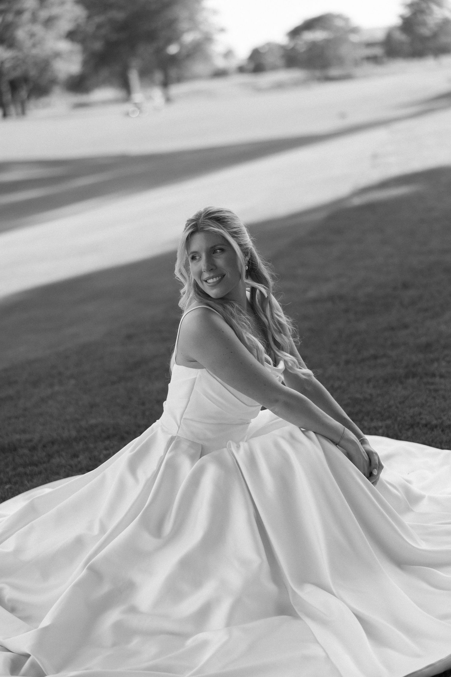 Black and white photo of smiling bride in ball gown sitting outdoors on wedding day.