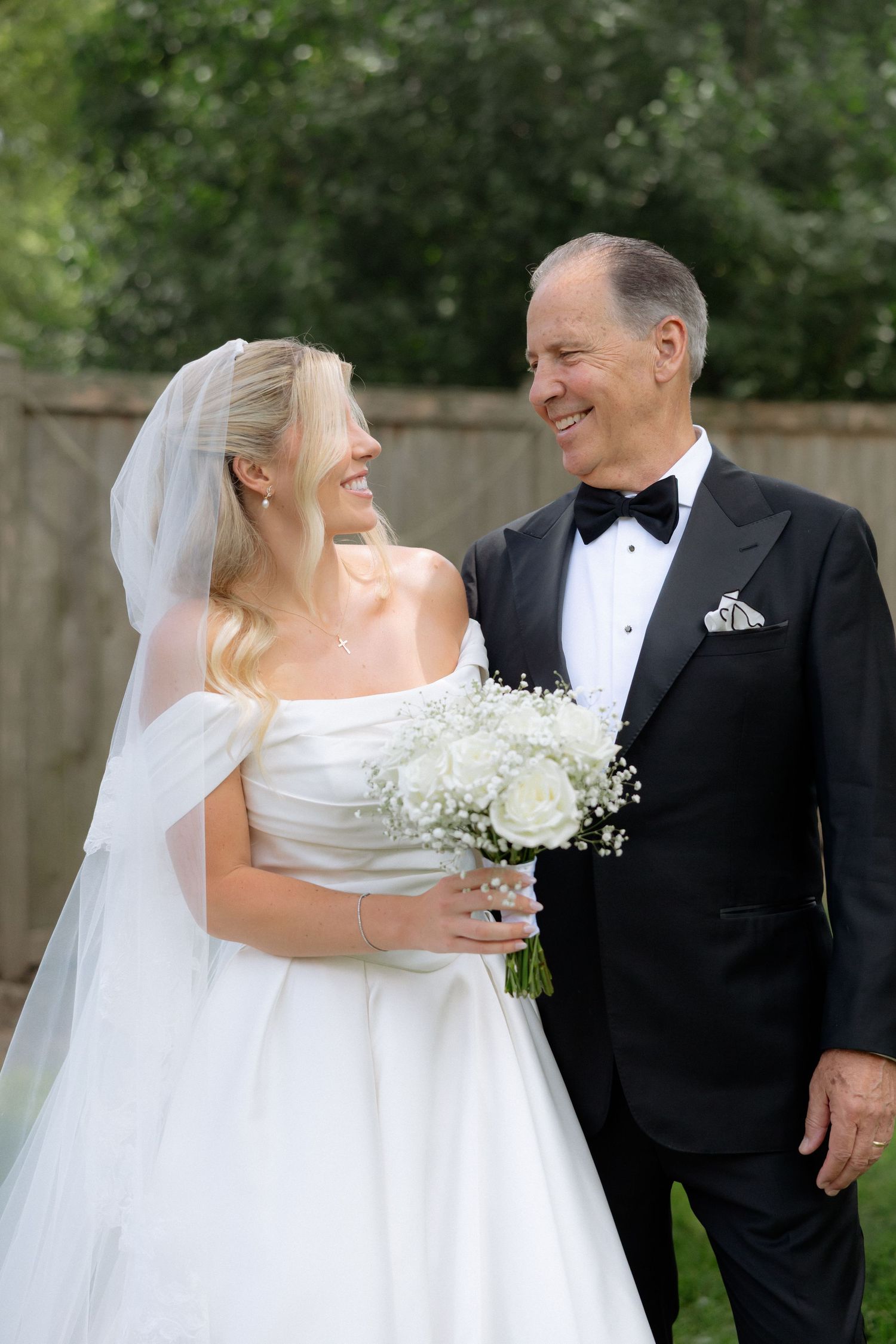 Bride in white off-shoulder gown and groom in tuxedo smile at each other in outdoor garden setting.