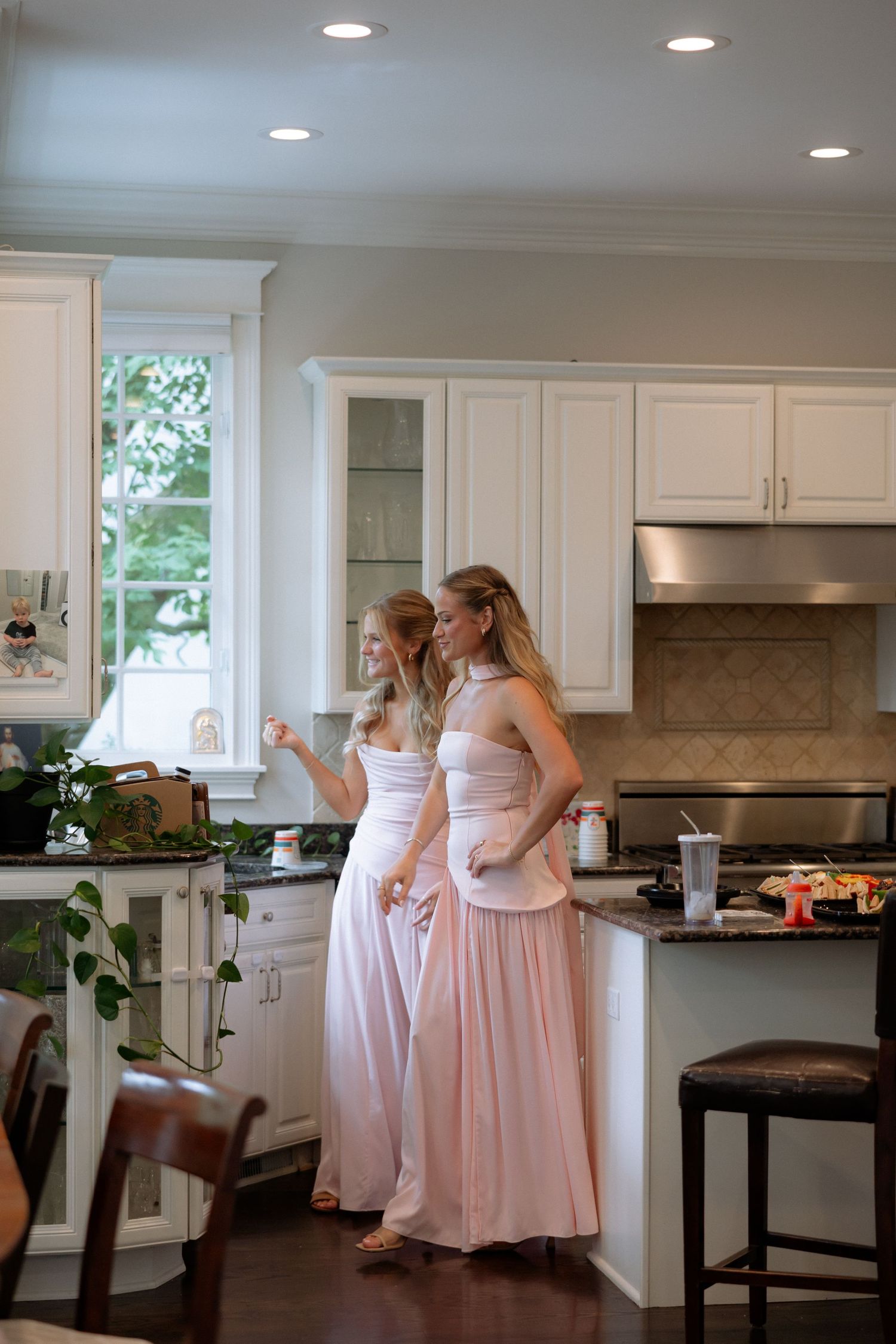 Two bridesmaids in blush pink gowns standing in a bright white kitchen before a wedding.