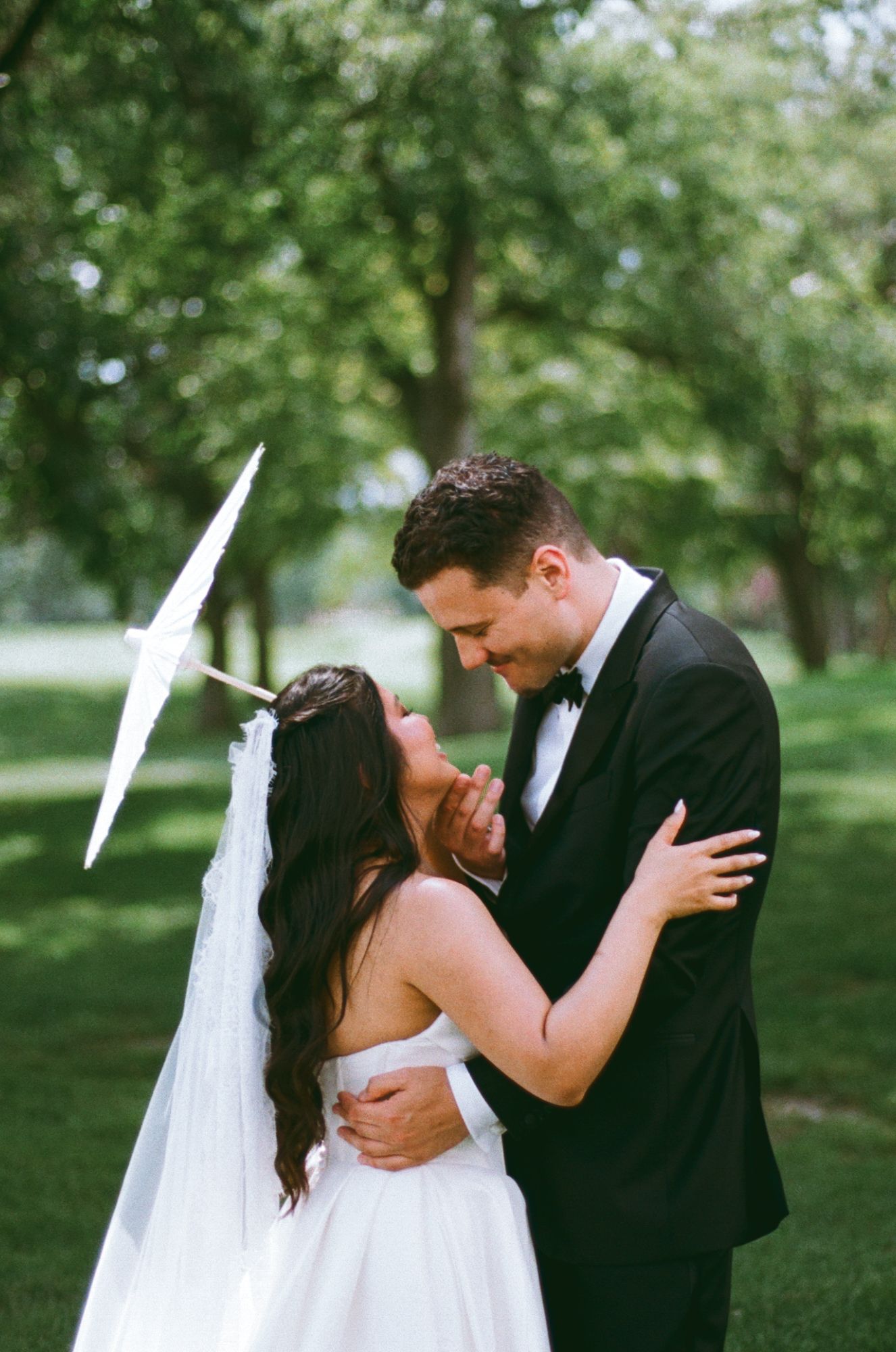 A bride and groom share a romantic moment outdoors, with the bride holding a white parasol and wearing a veil.