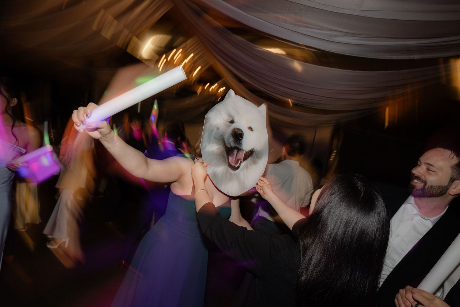 Party guests dance holding dog face sign and glow sticks at wedding.