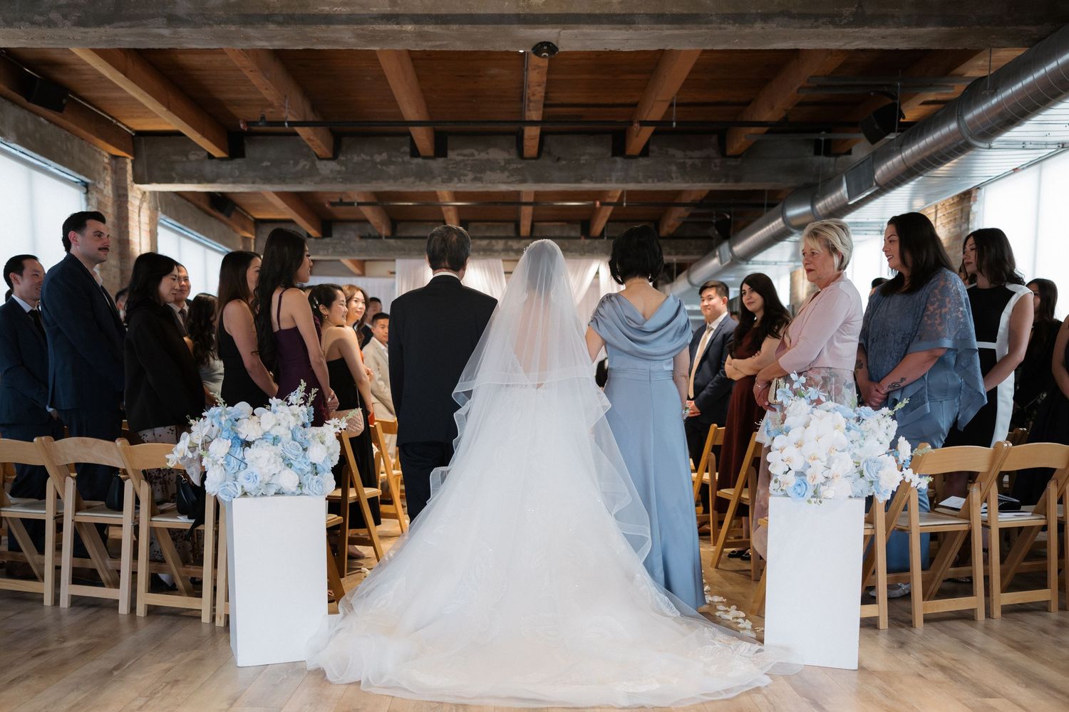 Bride in white gown and veil walks down aisle at elegant indoor wedding ceremony with blue floral arrangements.