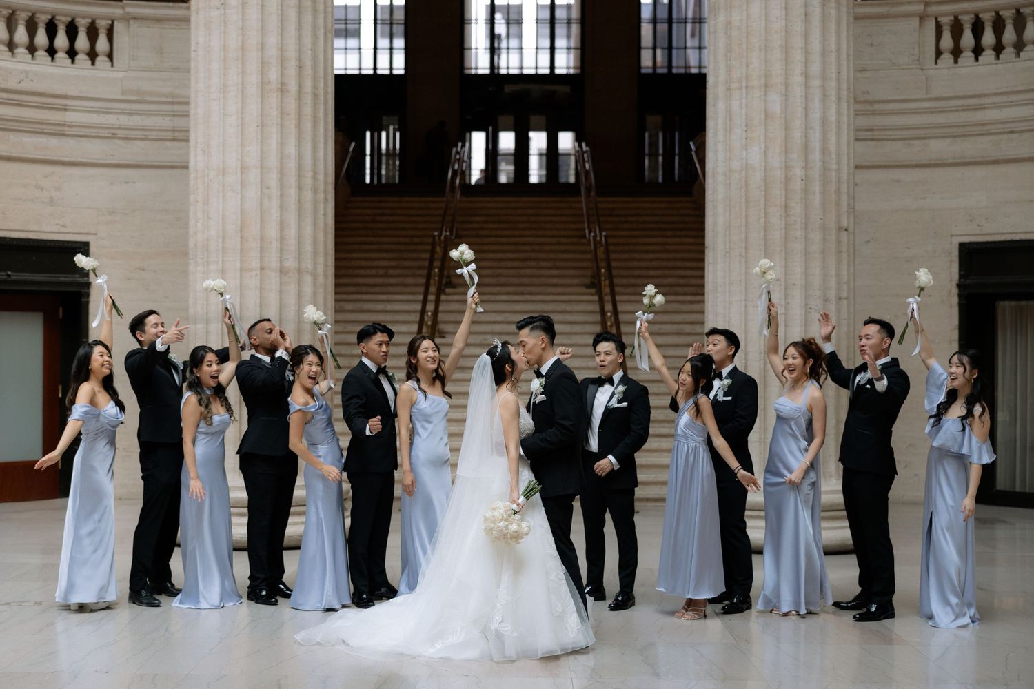 Bride and groom with wedding party in light blue dresses and black tuxedos raising bouquets.