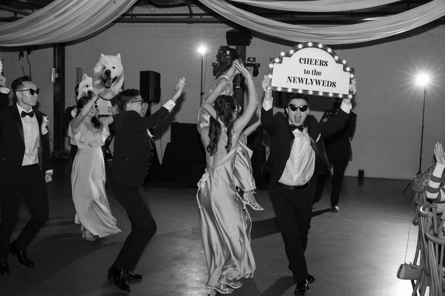 Black and white photo of wedding guests dancing joyfully at a reception with a Cheers to the Newlyweds marquee sign.