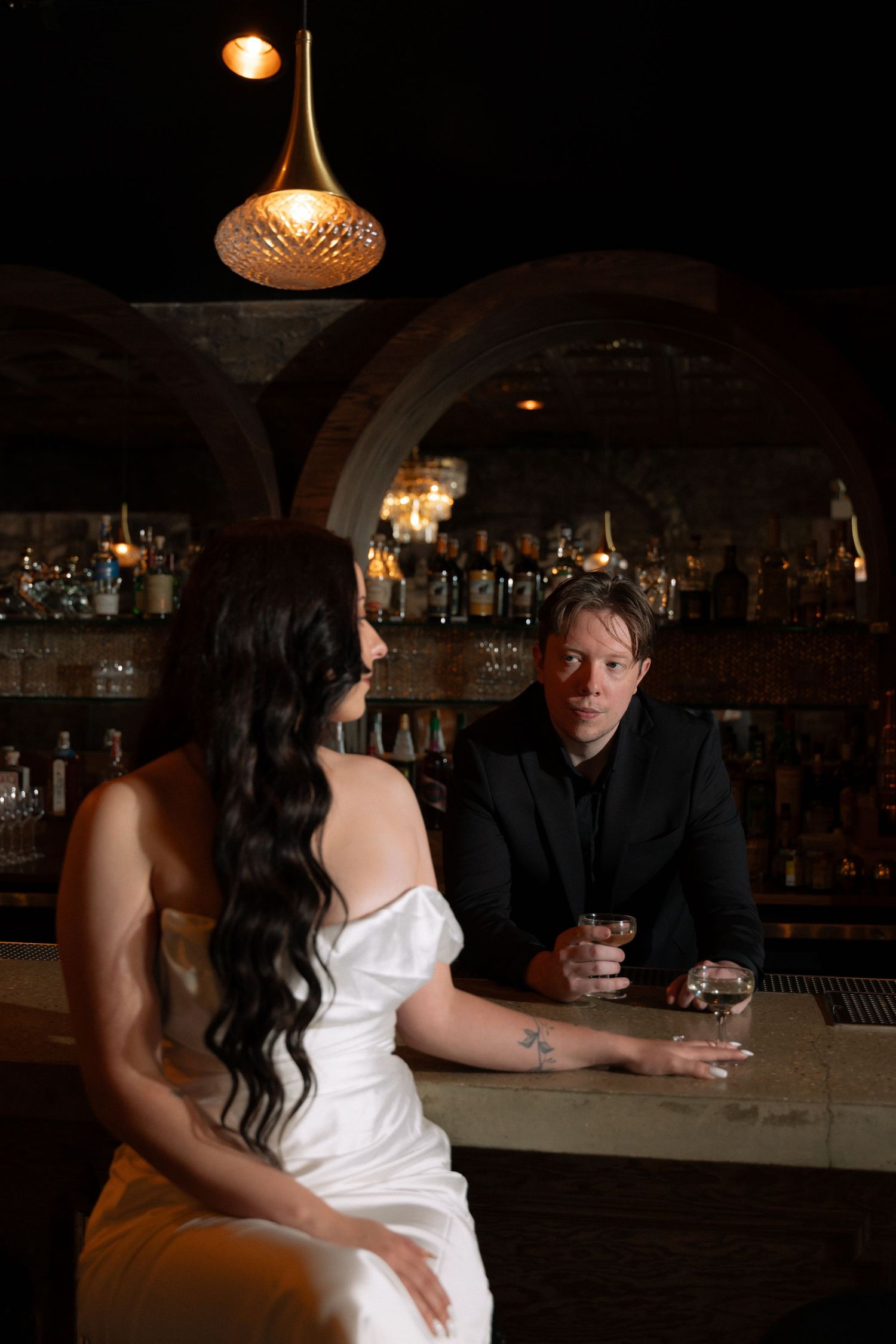 Bride in white satin gown and groom in black suit sharing drinks at a dimly lit upscale bar.