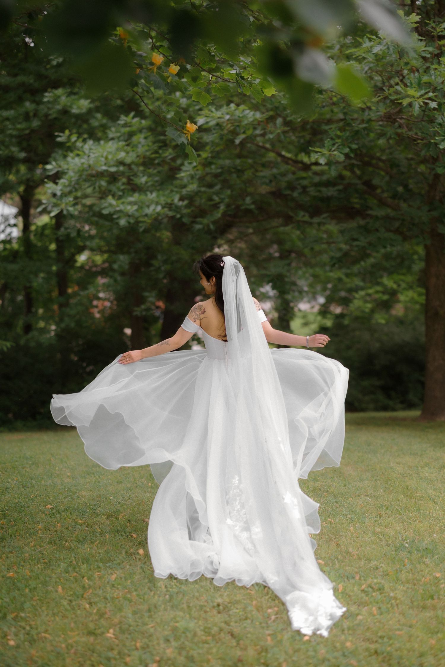 Bride in flowing white gown and veil twirling in lush green park, showcasing elegant lace wedding dress details.