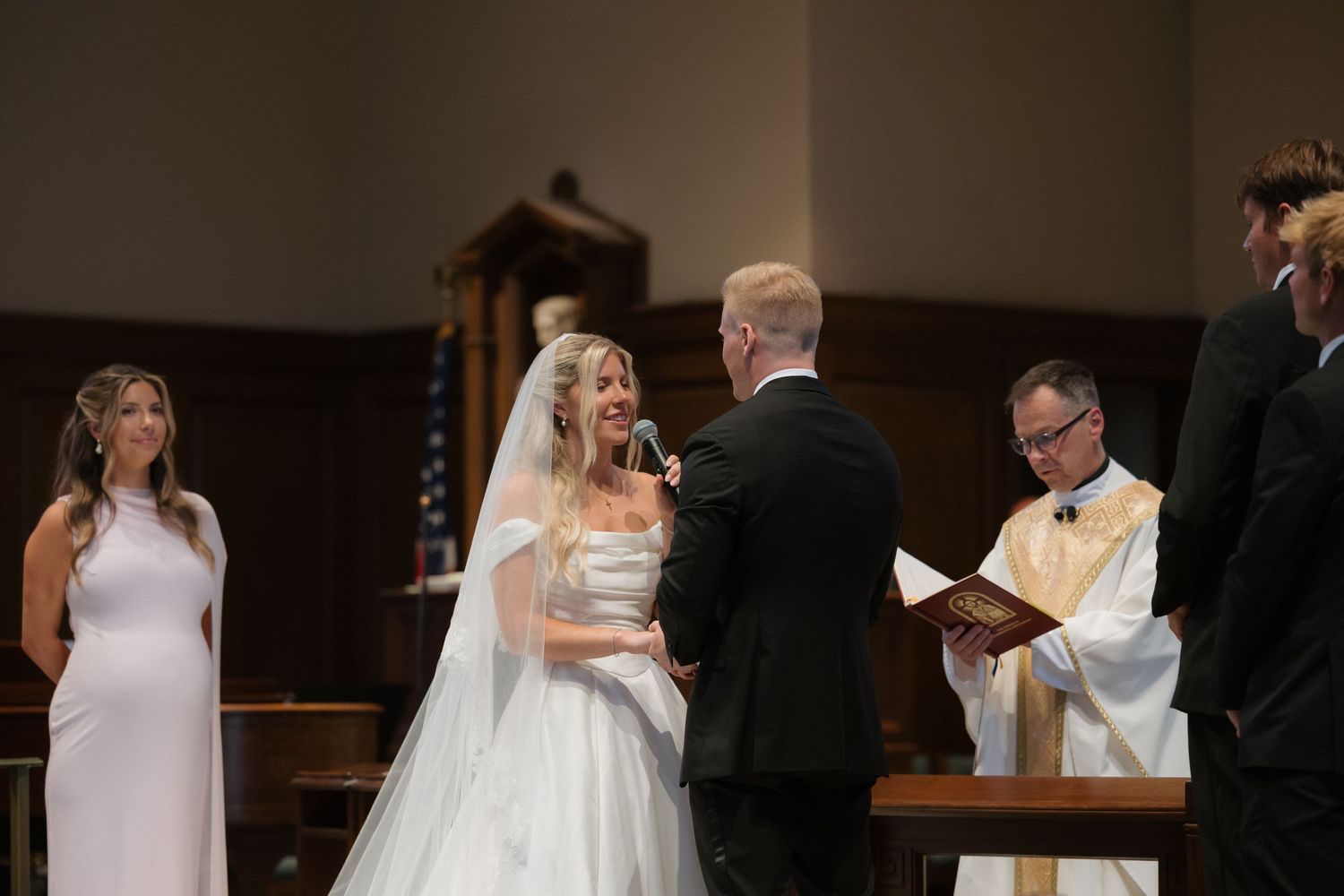 Bride speaks vows to groom at church altar with officiant and bridesmaid during wedding ceremony.