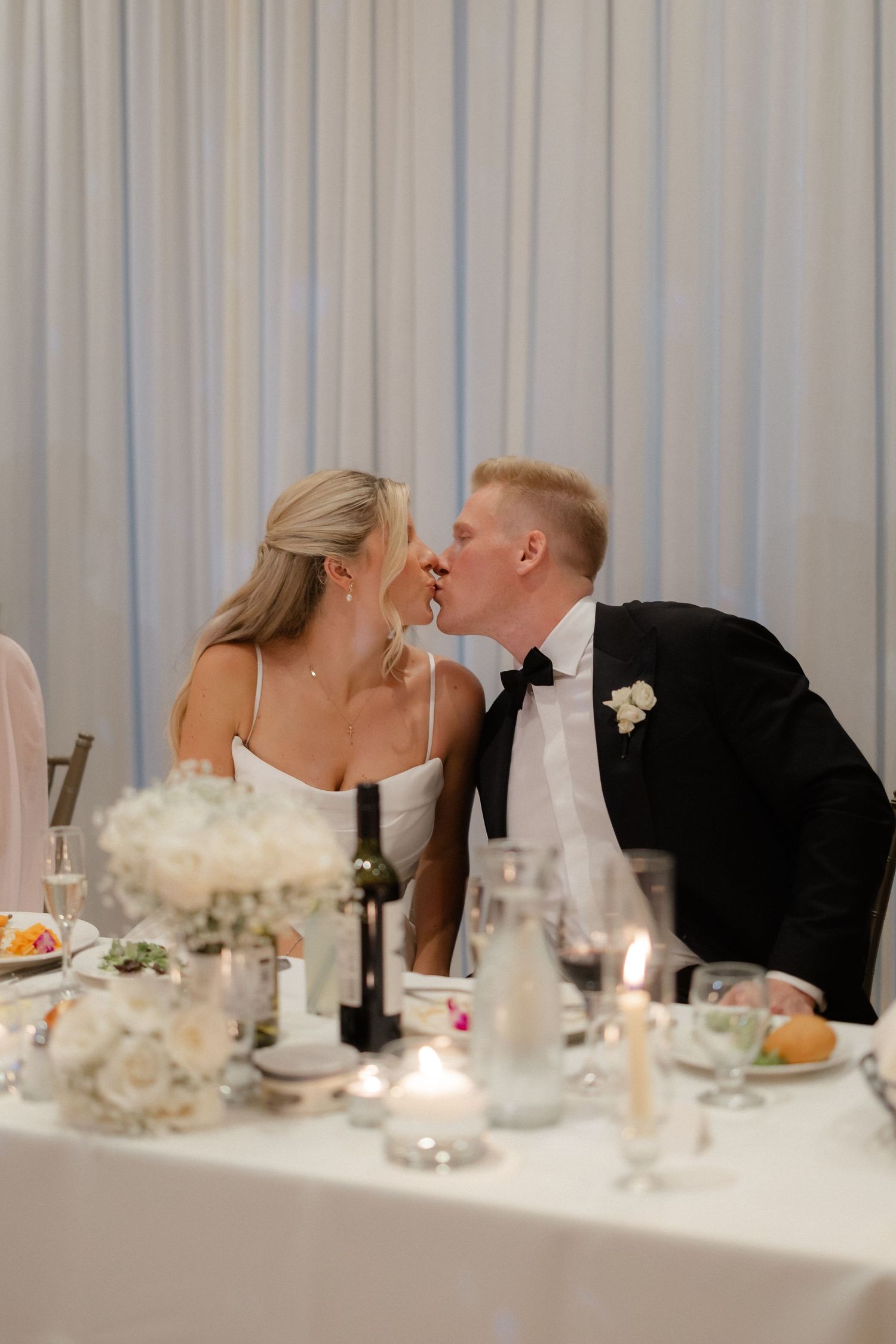 Bride and groom share a kiss at their wedding reception sweetheart table adorned with candles and white floral arrangements.