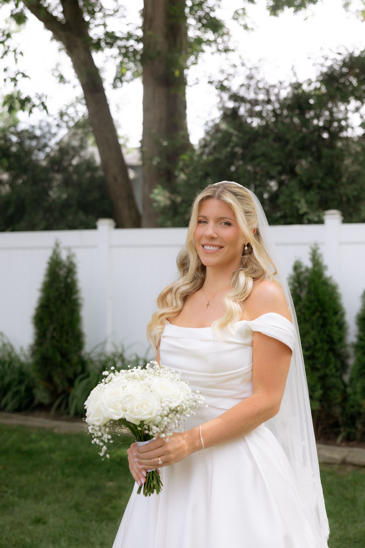 Smiling bride in off-shoulder white gown holds white rose bouquet outdoors near white fence and evergreen trees.