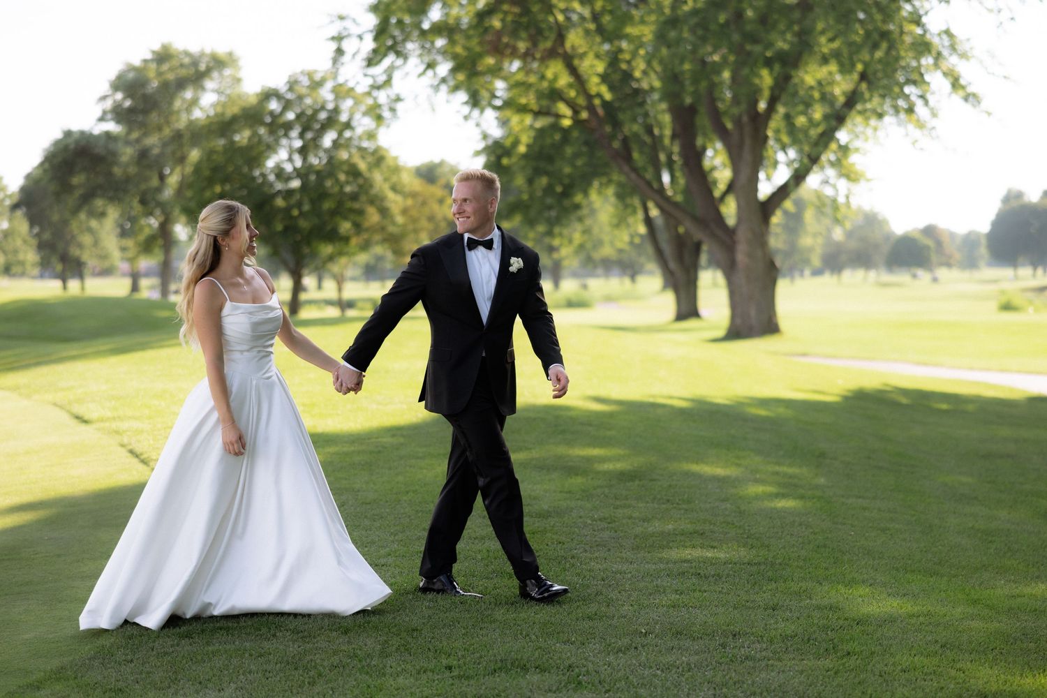 Bride and groom hold hands walking across lush green golf course on sunny wedding day.