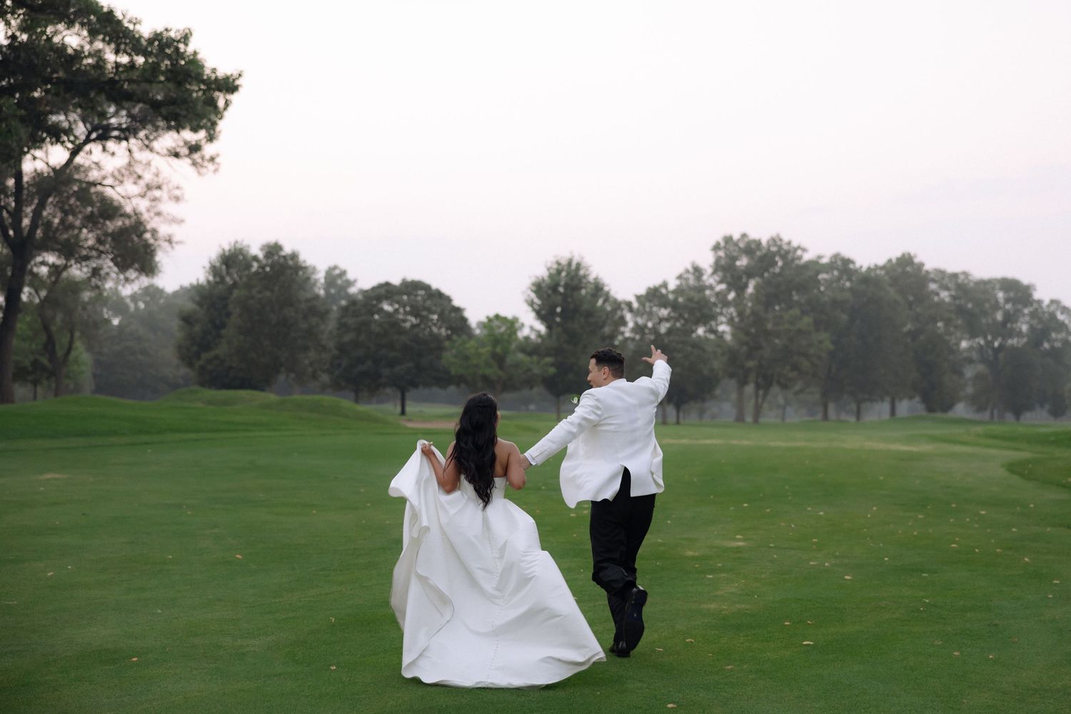 Bride in white gown and groom in white suit dancing on lush green golf course at dusk.