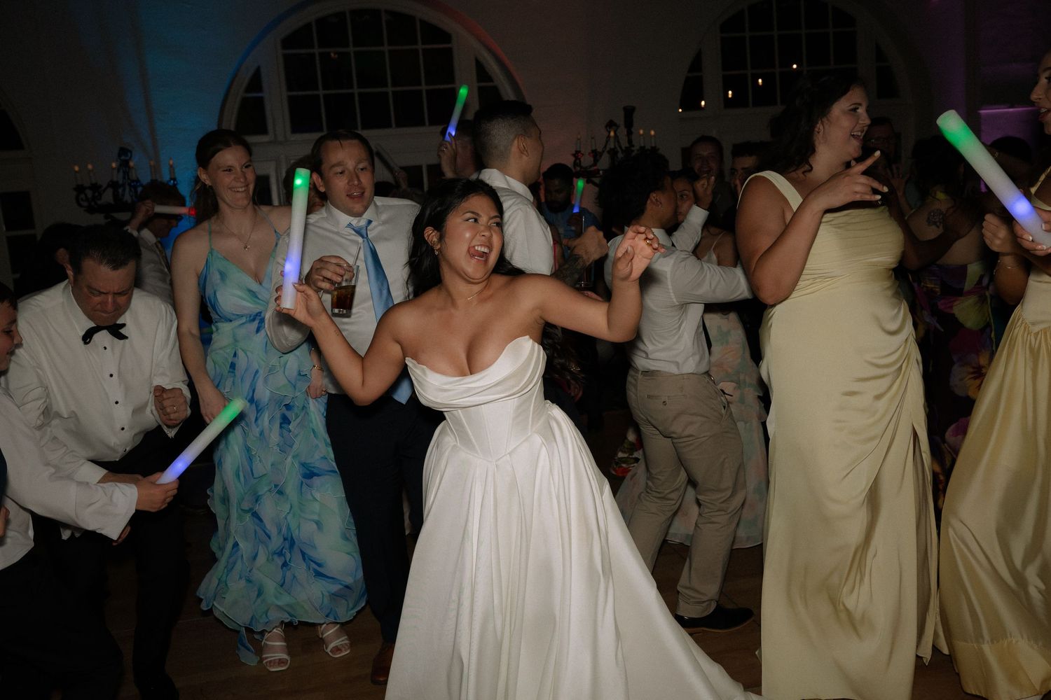 Bride dances enthusiastically with guests holding glow sticks at lively wedding reception party.