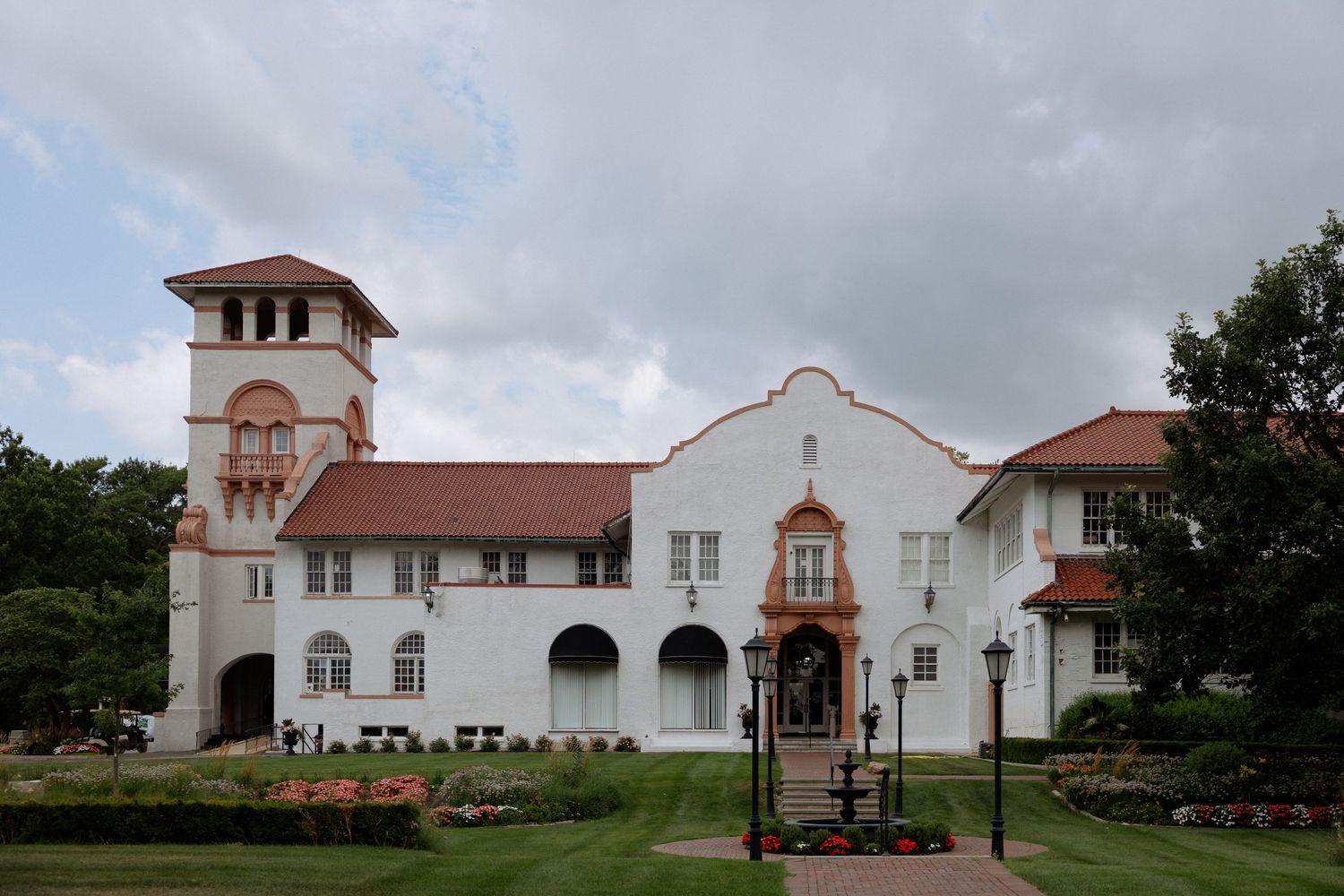 White Spanish Colonial Revival building with red tile roof, bell tower, and manicured gardens under a cloudy sky.