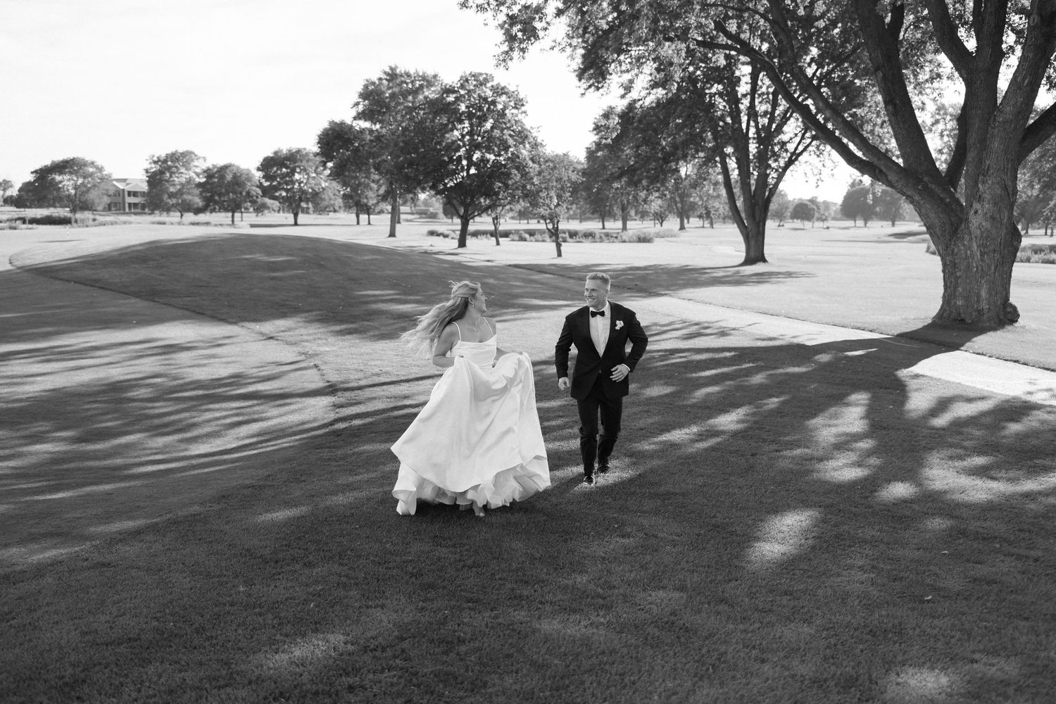Black and white photo of bride and groom walking hand in hand on a lush golf course path lined with trees.