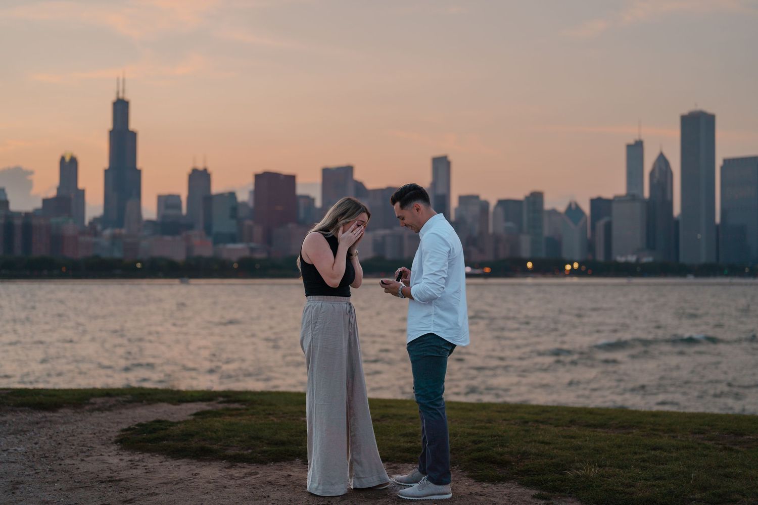 Man opening a ring box to propose to his girlfriend at sunset with the Chicago skyline behind them.