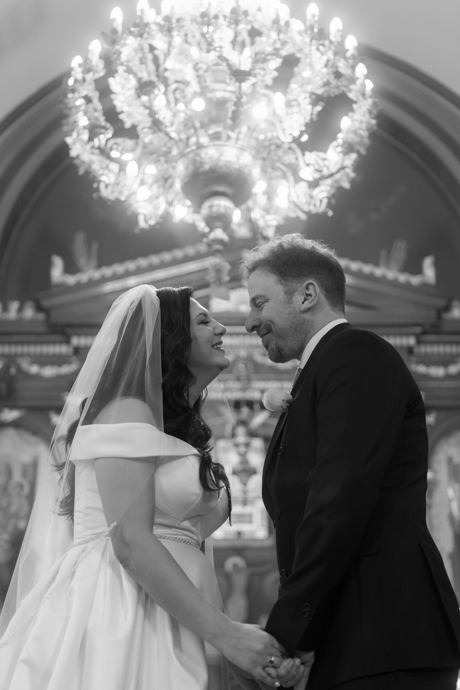 Black and white romantic portrait of bride and groom holding hands beneath grand chandelier in Orthodox church.