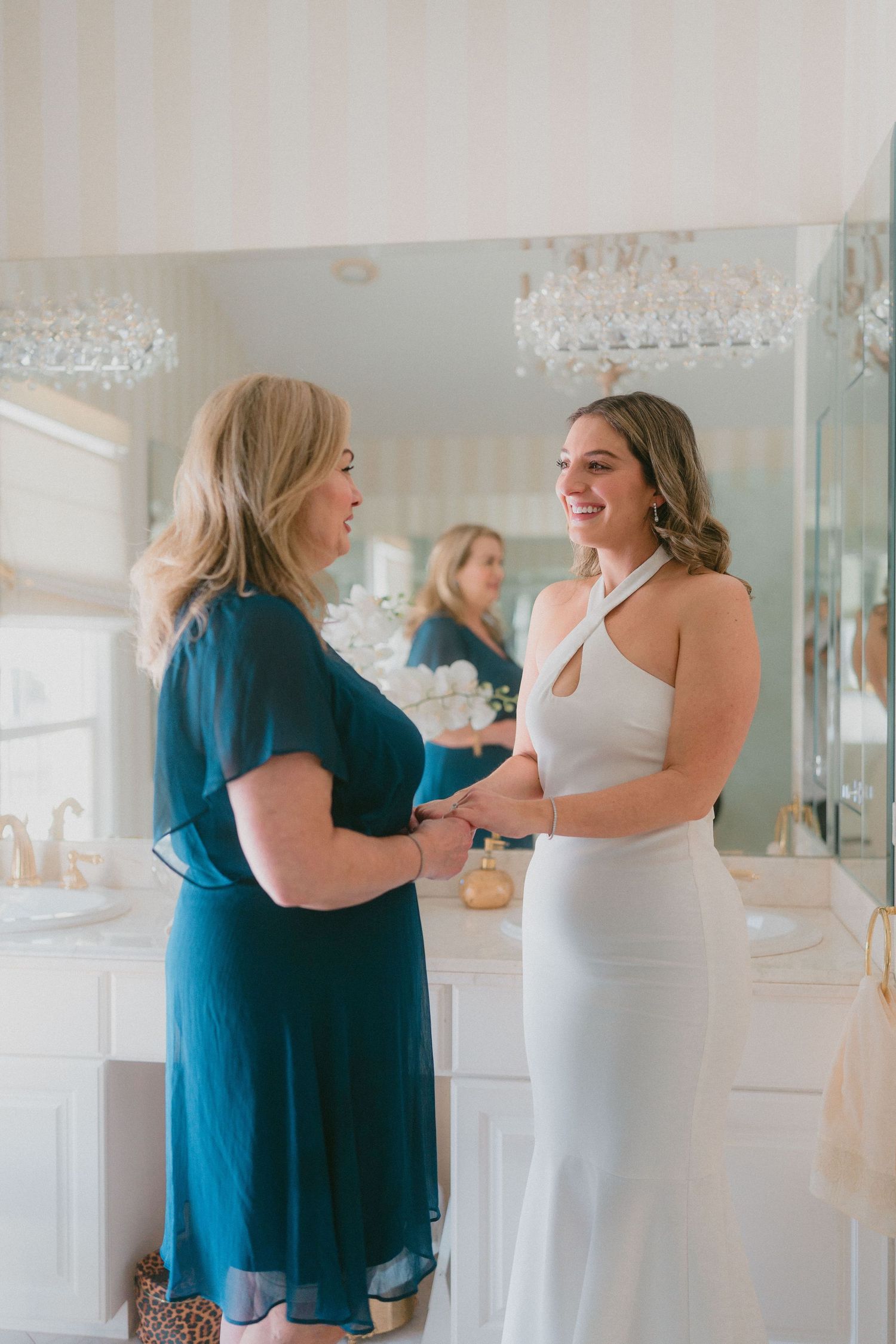 Bride in white halter gown smiling with older woman in teal dress in elegant bathroom setting.