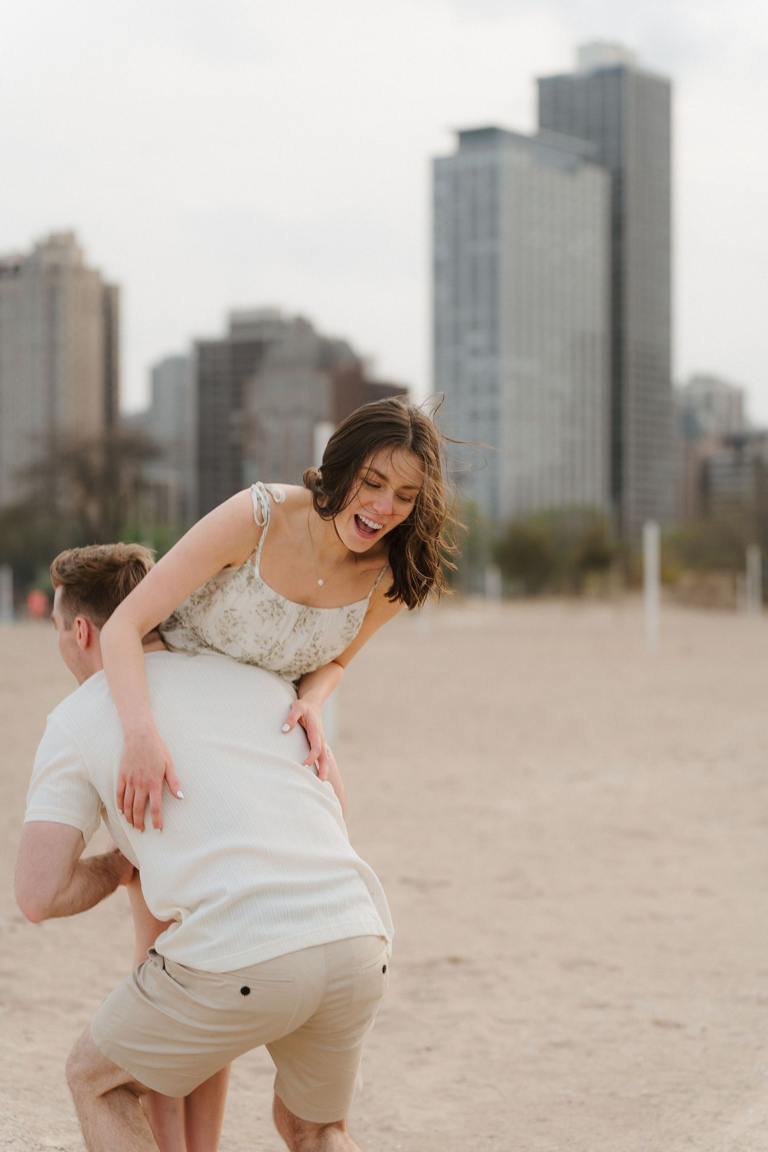 Laughing woman being lifted over a man's shoulder on a sandy beach with city skyscrapers in background.