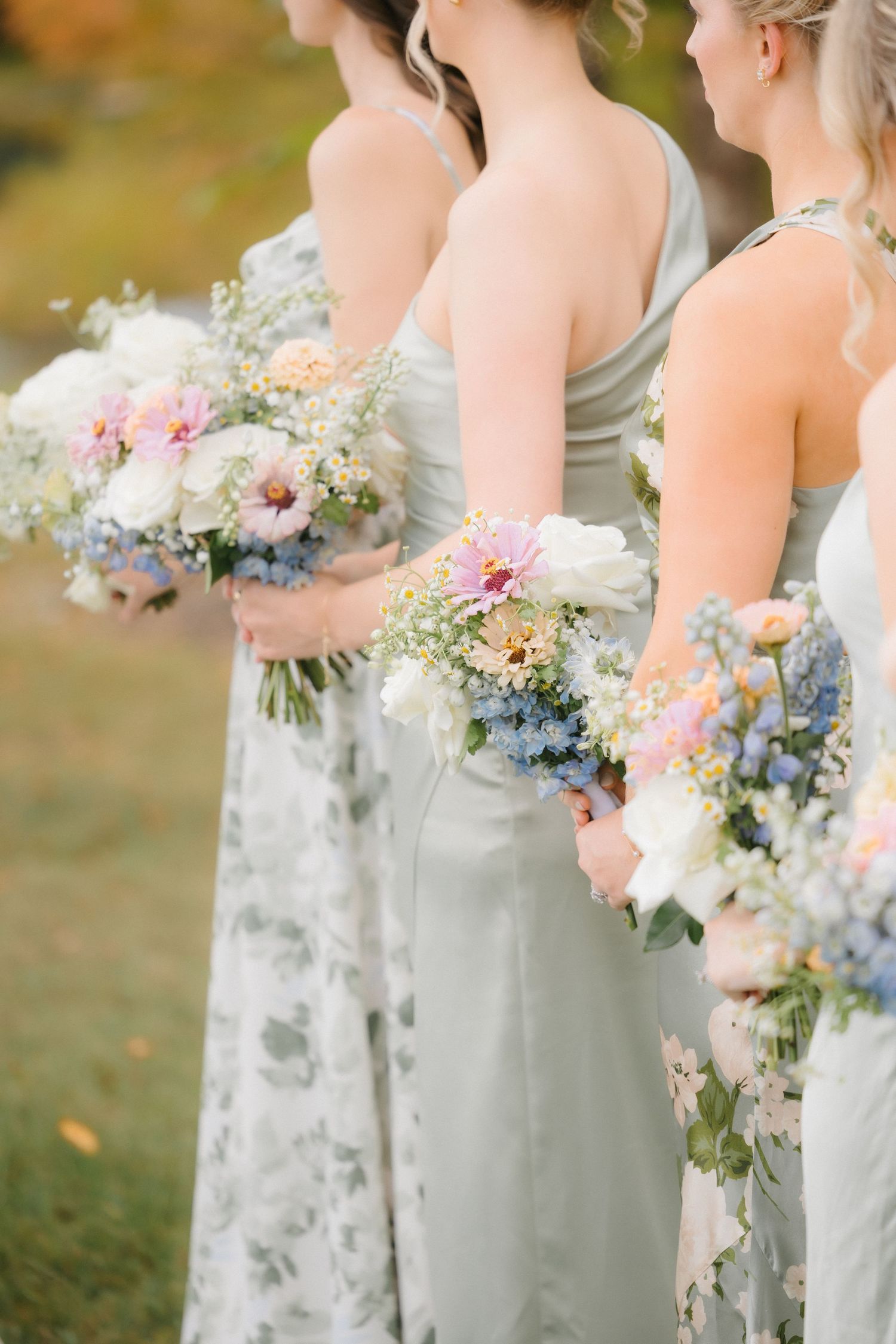 Three bridesmaids in sage green dresses hold colorful wildflower bouquets with pink blue and white blooms.