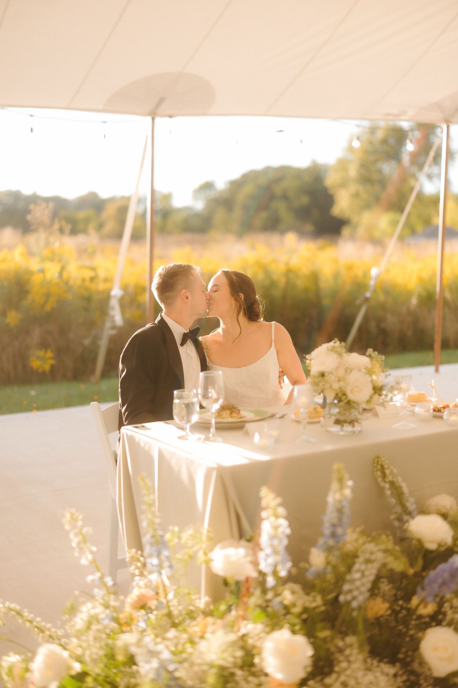 Newlyweds share a sweet kiss at their sweetheart table bathed in golden hour light with wildflowers in background.