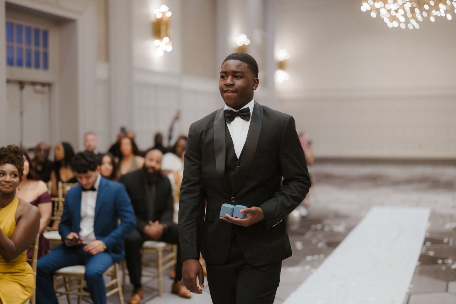 Young man in black tuxedo walks down wedding aisle in elegant venue with seated guests.