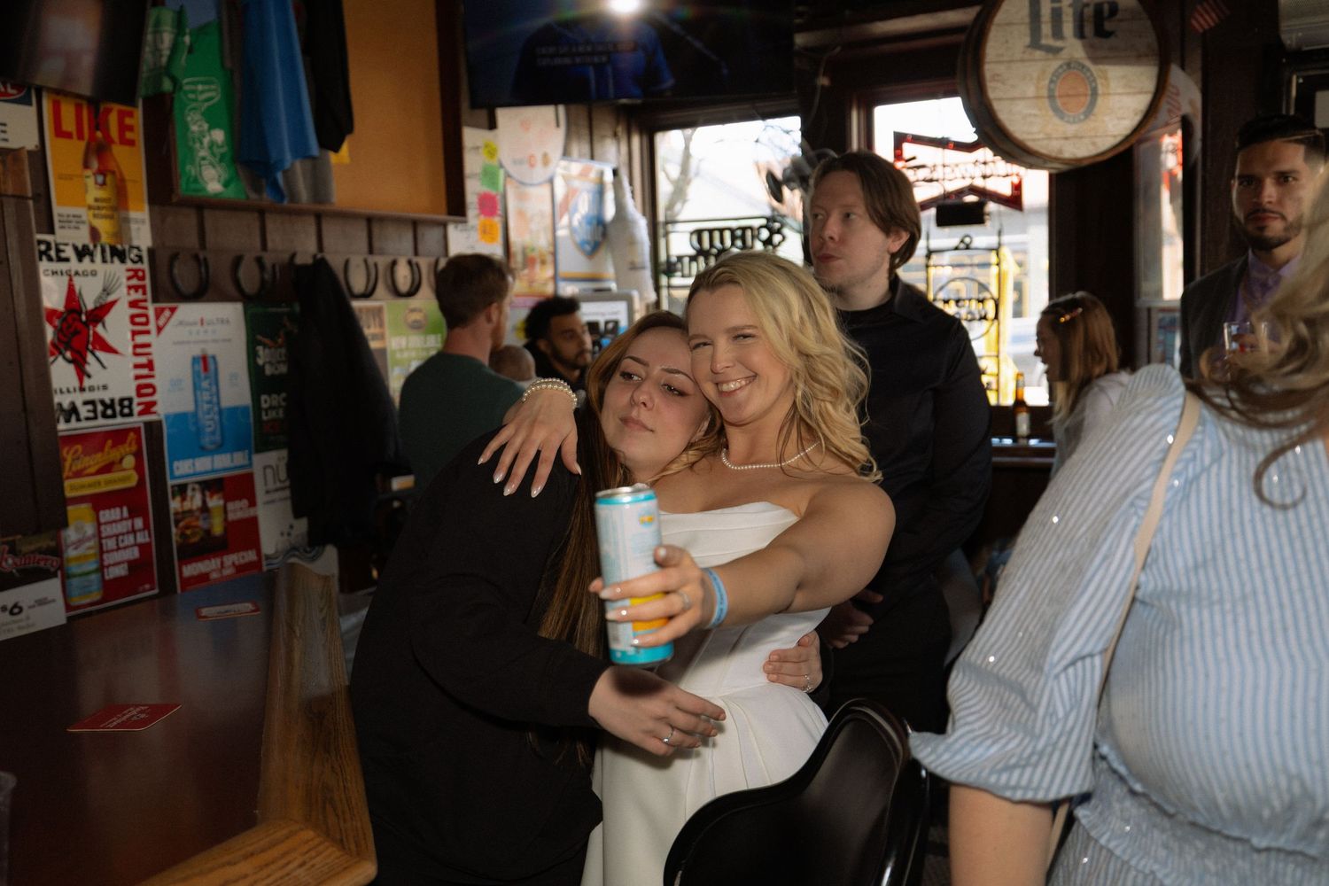 Two women hugging and smiling inside a lively bar, one holding a drink, with other patrons visible in the background.
