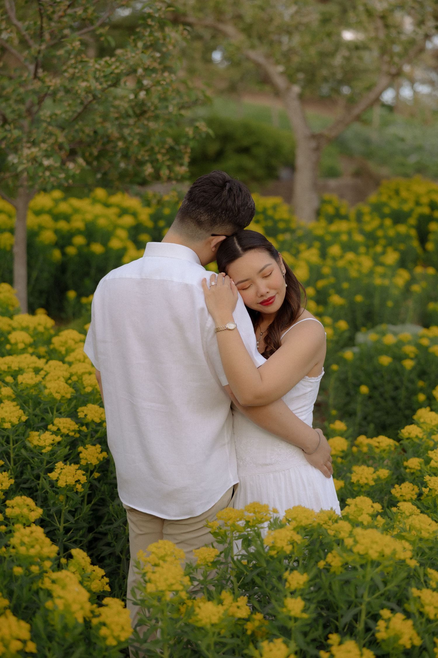 Couple embracing in a field of yellow flowers, woman in white dress resting her head on man's chest.