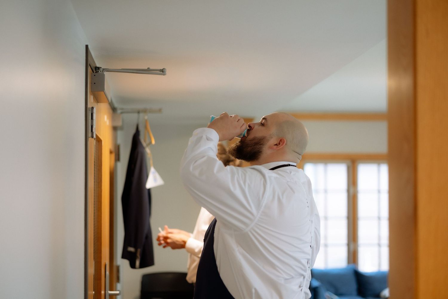 A bald groom in a white shirt adjusts his tie in a mirror while preparing for his wedding day.